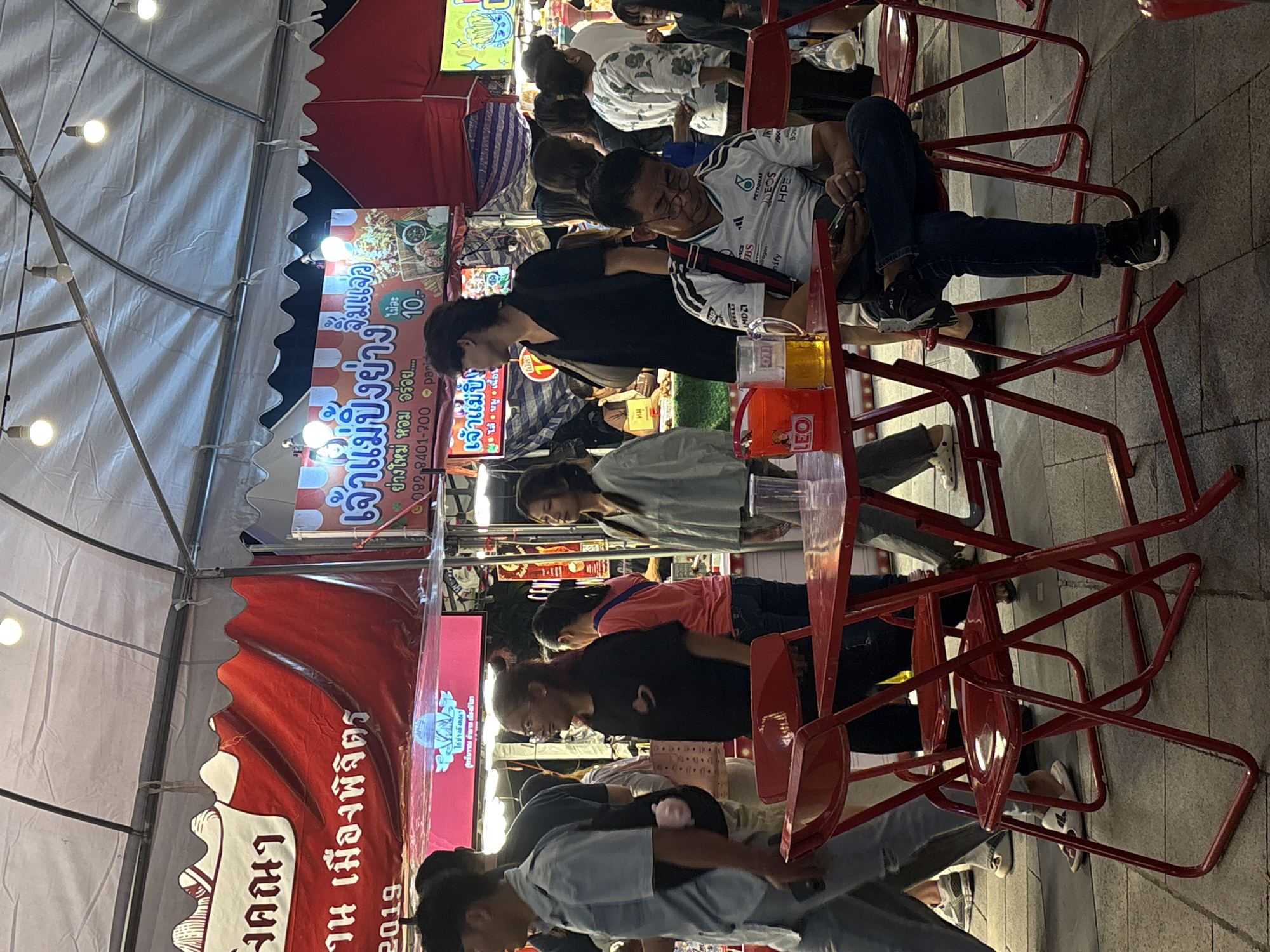 The image captures a bustling outdoor food market scene at night, likely in Chiang Mai City Municipality, Thailand, indicated by the Thai script visible on signage. The foreground features red metal tables and chairs, with a man sitting at one table, looking at his phone and wearing a white t-shirt with dark blue jeans. A glass of beer and a red bucket are on the table. Other patrons are visible in the background, some walking and others standing near food stalls. The stalls display colorful signs with Thai text, advertising various food items. Overhead, a white tent-like structure with lighting illuminates the area, suggesting a covered market. The ground is paved with cobblestones or similar material. The overall atmosphere is lively and typical of a night market in Thailand.
