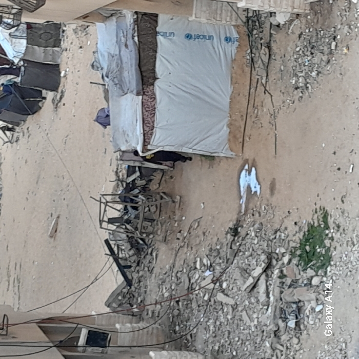 The image depicts a heavily damaged urban landscape repurposed into an informal settlement in Khan Yunis, Palestinian Territory. The ground consists primarily of sand and construction rubble, with large piles of concrete debris and twisted metal scattered throughout. In the mid-ground, a central dirt path, partially covered by scattered rubble, features a small, irregular puddle of liquid. To the left, a partially intact building of light-colored stone blocks has exposed wiring hanging from its exterior and what appears to be a dark rectangular monitor or panel mounted on its facade. Adjacent to this building, a significant mound of debris, including a skeletal metal frame, marks a destroyed structure. To the right of the path, a makeshift shelter is constructed from various materials, prominently featuring a white tarp with multiple blue "unicef" logos, topped by fabric and what appear to be old mattresses. A person in dark clothing is partially visible behind this shelter, moving from left to right. In the background, across a sandy expanse, several additional, more rudimentary makeshift tents or shelters, constructed from dark fabrics, are visible. A small patch of green vegetation grows amidst the rubble in the lower left foreground.