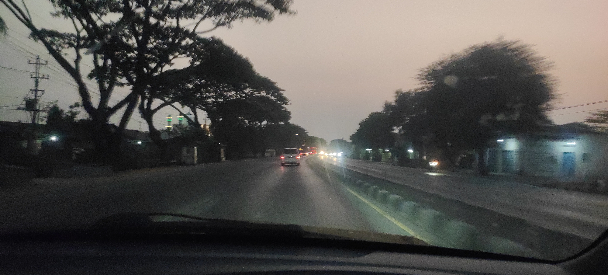 The image depicts a street scene in Cangkring Rembang, Indonesia, captured from the perspective of a moving vehicle. The sky is overcast and hazy, suggesting either early morning or late evening conditions, or possibly a dust-filled atmosphere. On the left side of the road, large trees with bare branches are silhouetted against the sky, with utility poles and wires visible among them. Further down the road on the left, there are some low-lying structures with lights on, and a building with green and yellow illuminated domes, likely a mosque, is visible in the distance.
The road itself is wet and reflective, indicating recent rain or a damp surface, with yellow lines marking the lanes. Several vehicles are on the road, including a white van in the foreground and other cars further ahead, their headlights illuminated. The right side of the road features more trees and some buildings, also with lights on. A concrete barrier with rounded protrusions separates the lanes in the distance. The overall impression is one of a somewhat busy street in a semi-urban or rural area, likely during twilight hours. The image quality is somewhat blurred, consistent with being taken from a moving vehicle.