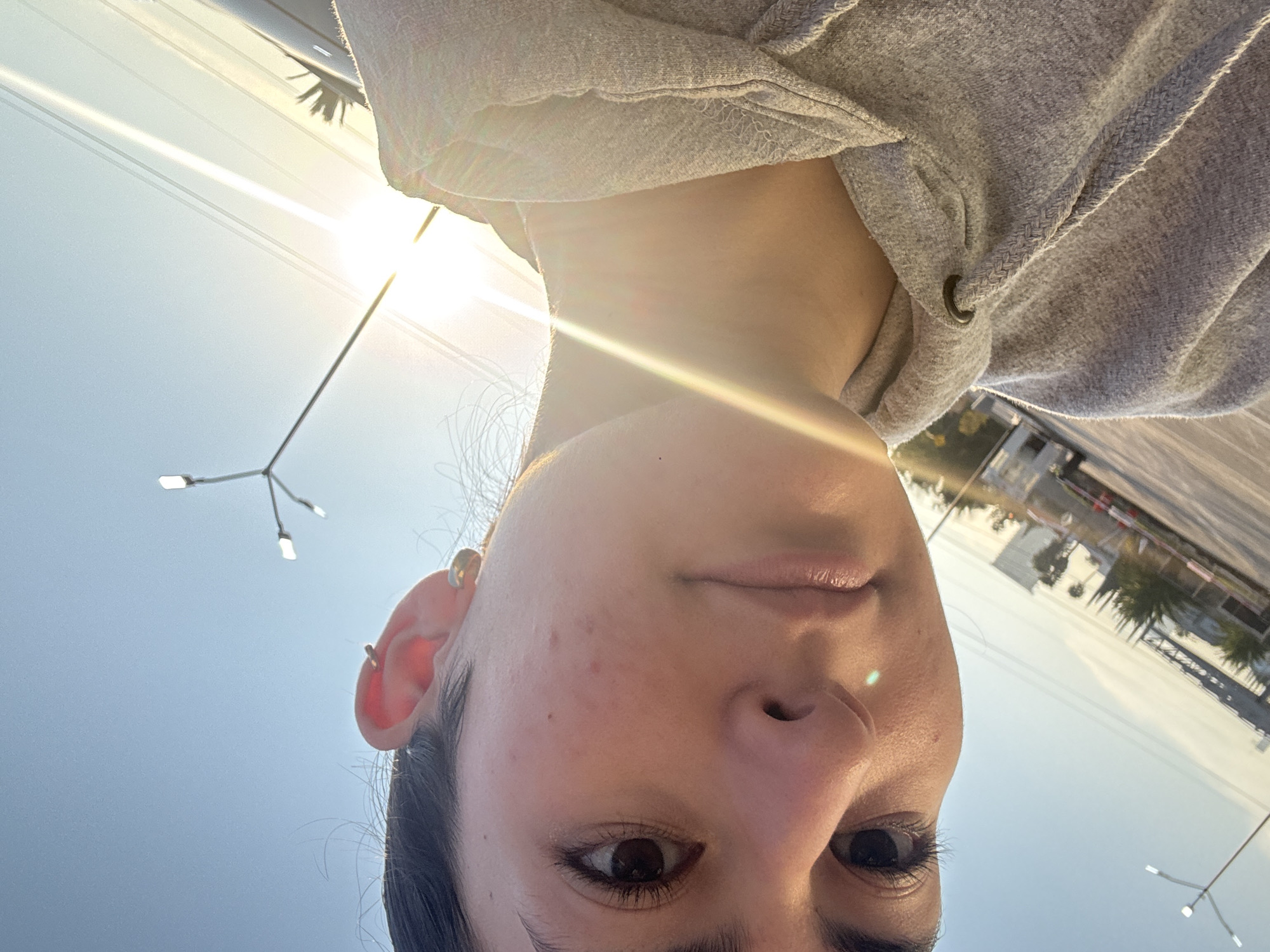 A selfie was taken in San Bernardino Tlaxcalancingo, Mexico, on what appears to be a clear, sunny day. The subject, a person with fair skin, dark hair, and brown eyes, is wearing a gray hooded sweatshirt. The head is angled downward, with the face taking up most of the frame. A streetlight is visible in the background. In the far background are buildings, trees, and a road.