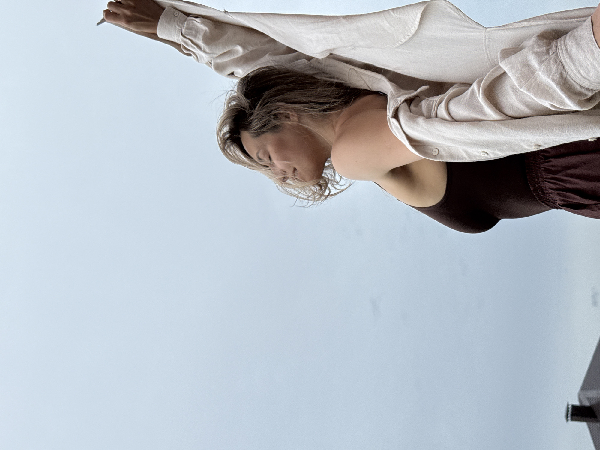 A woman with blonde hair, wearing a brown tank top and an unbuttoned beige shirt, is pictured from the chest up, with her head tilted back and eyes closed. She is reaching her right arm upwards, holding a pen or pencil. The background is a pale blue sky with some subtle clouds, and a hint of a house roofline is visible in the lower left corner, possibly indicating a suburban or semi-rural setting within or near Chaikovsky, Russia. The lighting suggests an overcast day.