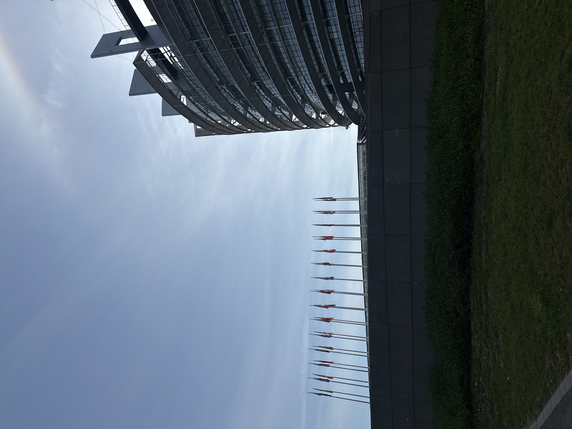 The image displays a modern architectural landscape in Strasbourg, France, captured during bright daylight. The main subject is a large, striking building, likely the European Parliament, which dominates the upper left portion of the frame. Its facade features a distinctive, curved design with a grid-like structure of metal and glass, giving it a contemporary and imposing appearance.
Below the building, a long line of flagpoles stands prominently, flying numerous national flags, which, given the context, are likely those of European countries. These flags are set against a vast expanse of bright blue sky, which fills a significant portion of the image. The sky itself is mostly clear with a few faint clouds, and a subtle rainbow-like halo or atmospheric optical phenomenon is visible in the upper left corner.
To the right of the flags and the building's base, a dark, tiled or paved area transitions into a neat strip of vibrant green grass, hinting at landscaped grounds surrounding the structure. No people or specific activities are visible, and there is no discernible text within the image. The scene conveys a sense of institutional grandeur and modernity, under clear, bright weather conditions.