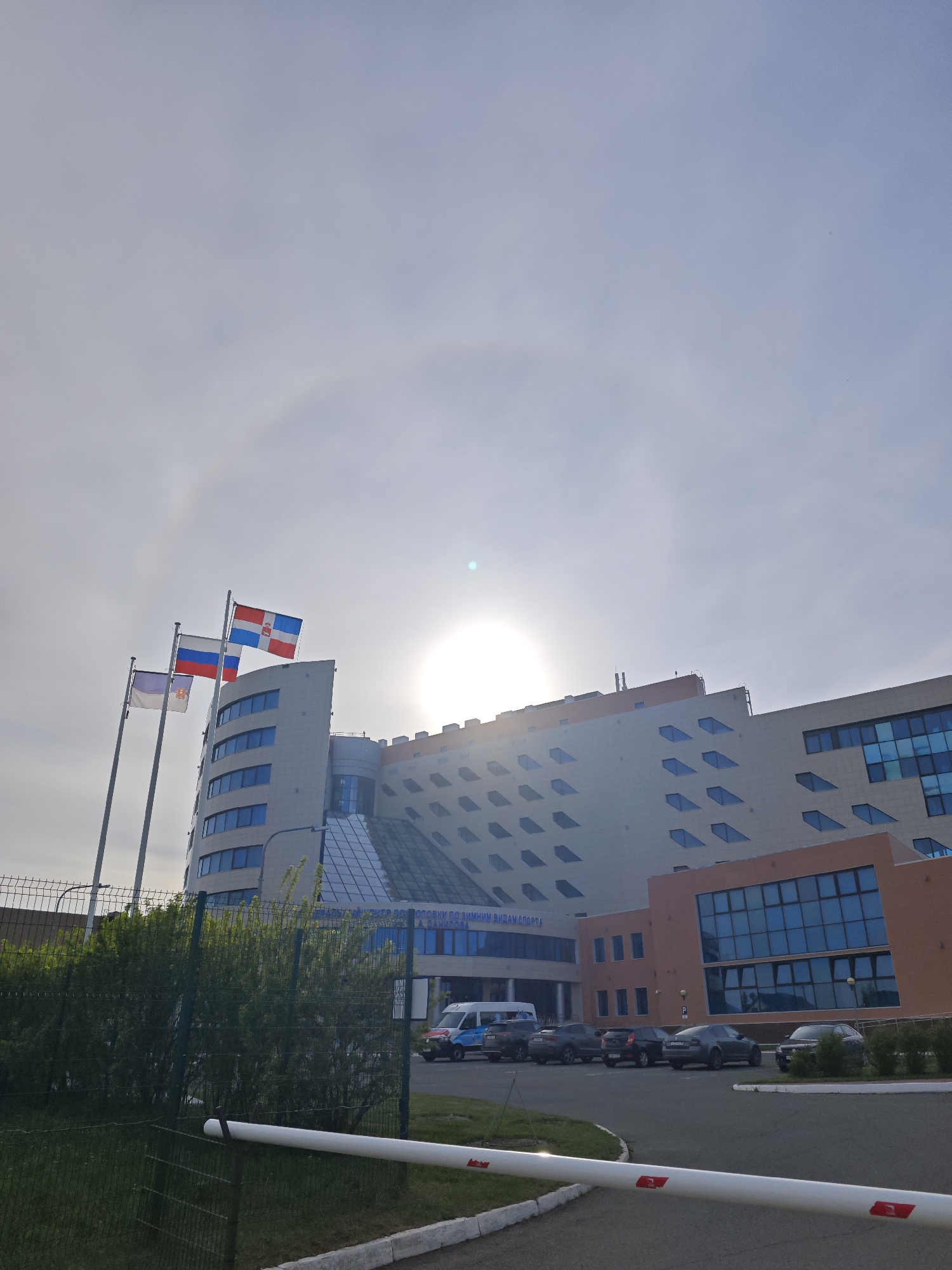 The image shows a multi-story building complex in Прикамский, Russia, with three flags flying in front of it. The flags are, from left to right, a white flag with a blue cross, the Russian flag, and a red, white, and blue flag. The building has a modern design with blue-tinted windows. A security fence and a gate are in the foreground. Several cars are parked near the building's entrance. A halo is visible around the sun in the sky.