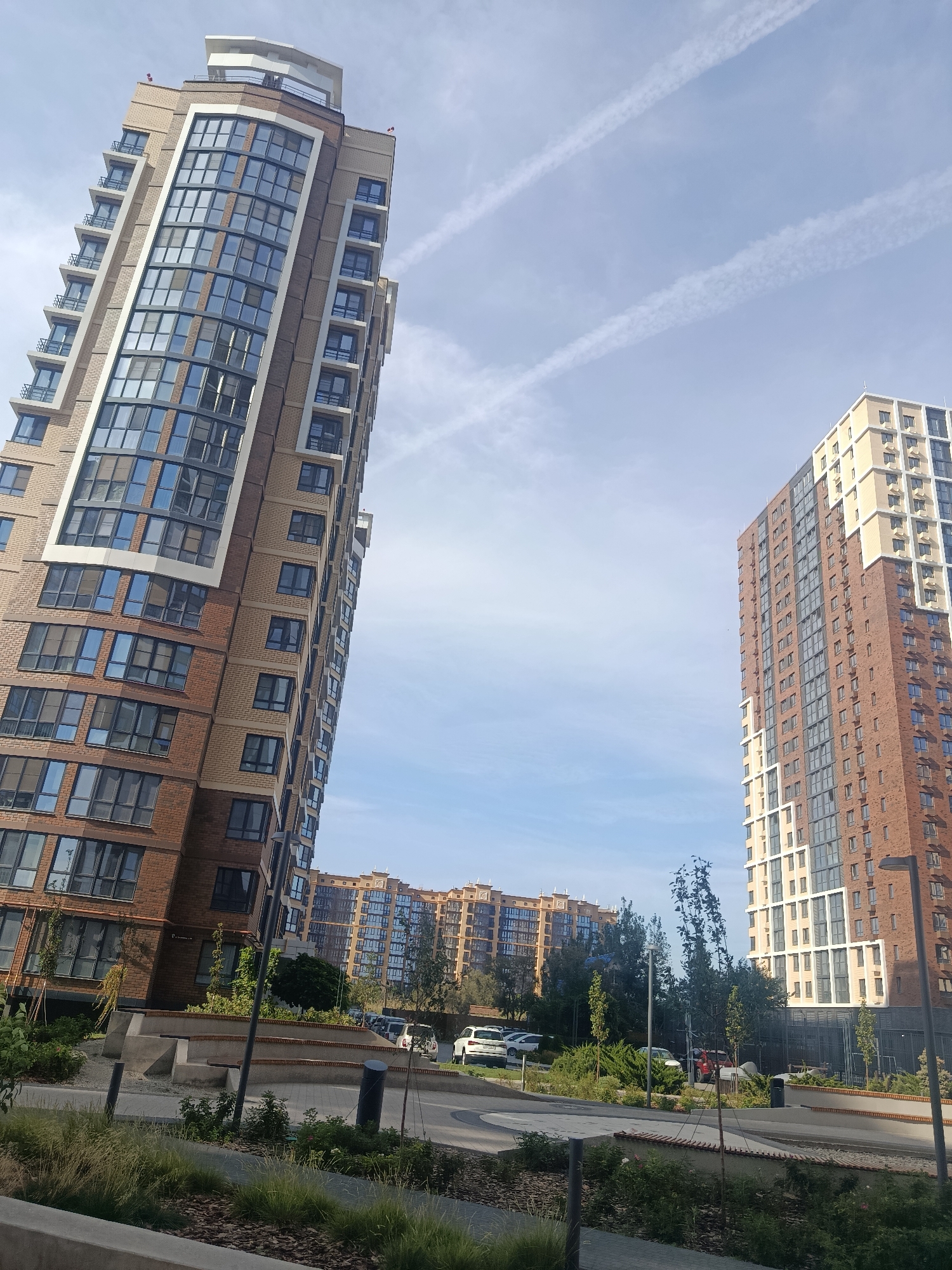 The image depicts a modern residential complex with multiple tall buildings. The architecture features brickwork and large glass windows, with balconies on many levels. In the foreground, there is a landscaped area with grass and shrubs, as well as paved pathways. Several cars are parked in the background, suggesting this is a residential area. The sky is blue with some scattered clouds and contrails from airplanes. The overall impression is one of a contemporary urban development. There is no visible indication of the city of Astrakhan, Russia, in the image.