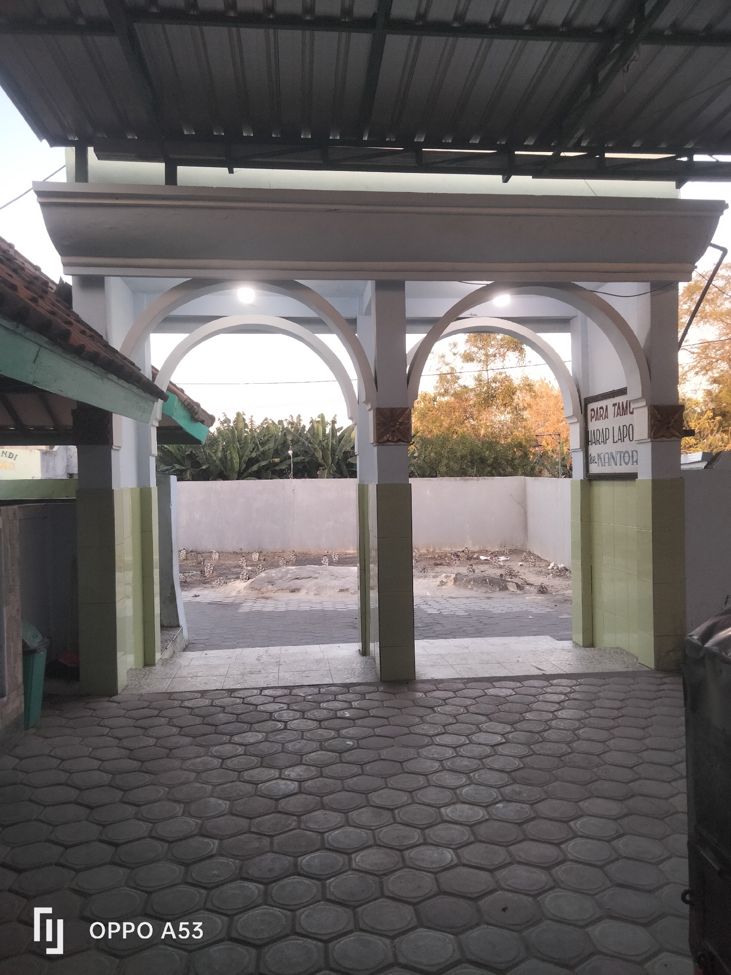 The image captures an arched entryway, possibly at a building in Badung, Indonesia, given the text on a sign. The entryway features two large arches supported by pillars. A metal roof extends over the entryway. To the left, a section of a building with a tiled roof is visible, along with a green trash can. The ground in front of the entryway is paved with hexagonal stones. Beyond the arches, a concrete wall encloses an area with sparse vegetation and debris. A sign mounted on a pillar reads "PARA TAMU HARAP LAPO ke KANTOR", which translates to "GUESTS PLEASE REPORT TO THE OFFICE". The lighting suggests it is either early morning or late afternoon. No people are visible in the scene.