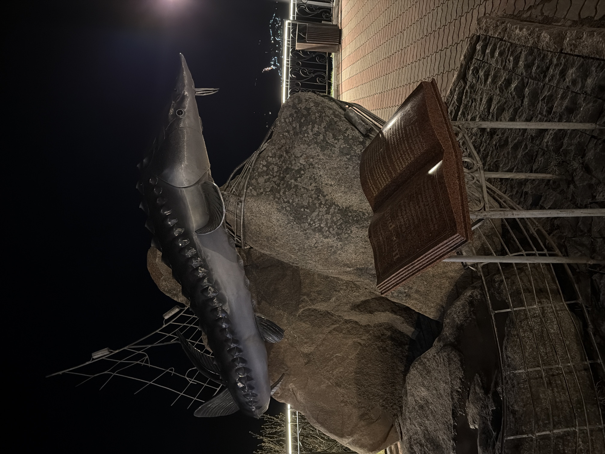 The image shows a monument in Слизнево, Russia, featuring a large metal sturgeon sculpture vertically mounted next to a stone structure and an open book with Cyrillic text. The monument is situated outdoors, likely in a public area, with a brick walkway visible in the background. The scene is illuminated by artificial lighting, and a metal fence is present.