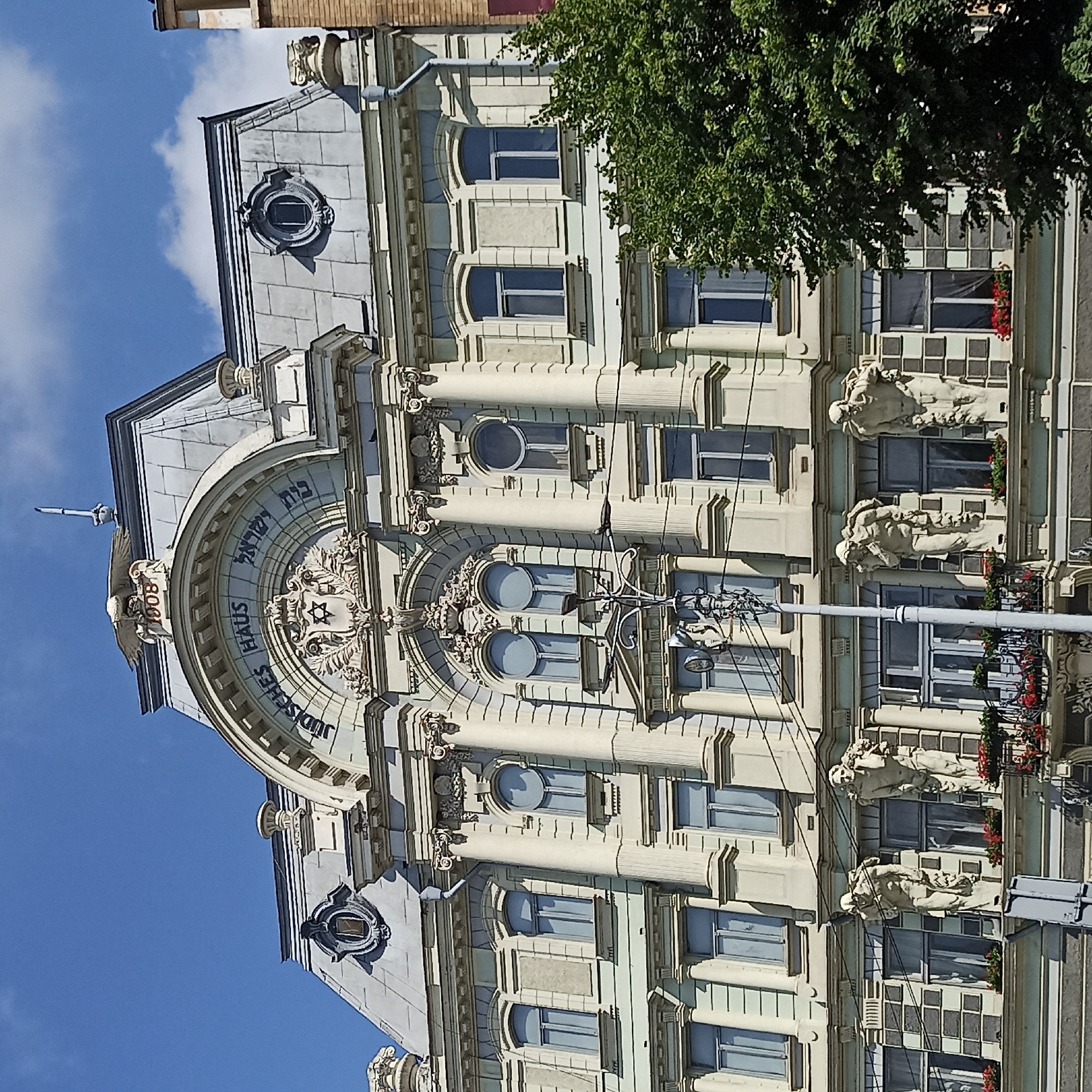 The image shows the "Jewish House" building in Chernivtsi, Ukraine, a historic architectural landmark. The building features ornate classical architecture with columns, decorative carvings, and a mansard roof. Above the entrance arch, the inscription "JUDISCHES HAUS" (Jewish House) is visible, along with the year "1908" and Hebrew inscriptions. Two statues of standing figures adorn the facade, and flowering plants are visible on balconies. The sky is blue with some clouds, and a green tree is partially visible on the right side of the image. Electrical wires are strung across the facade.