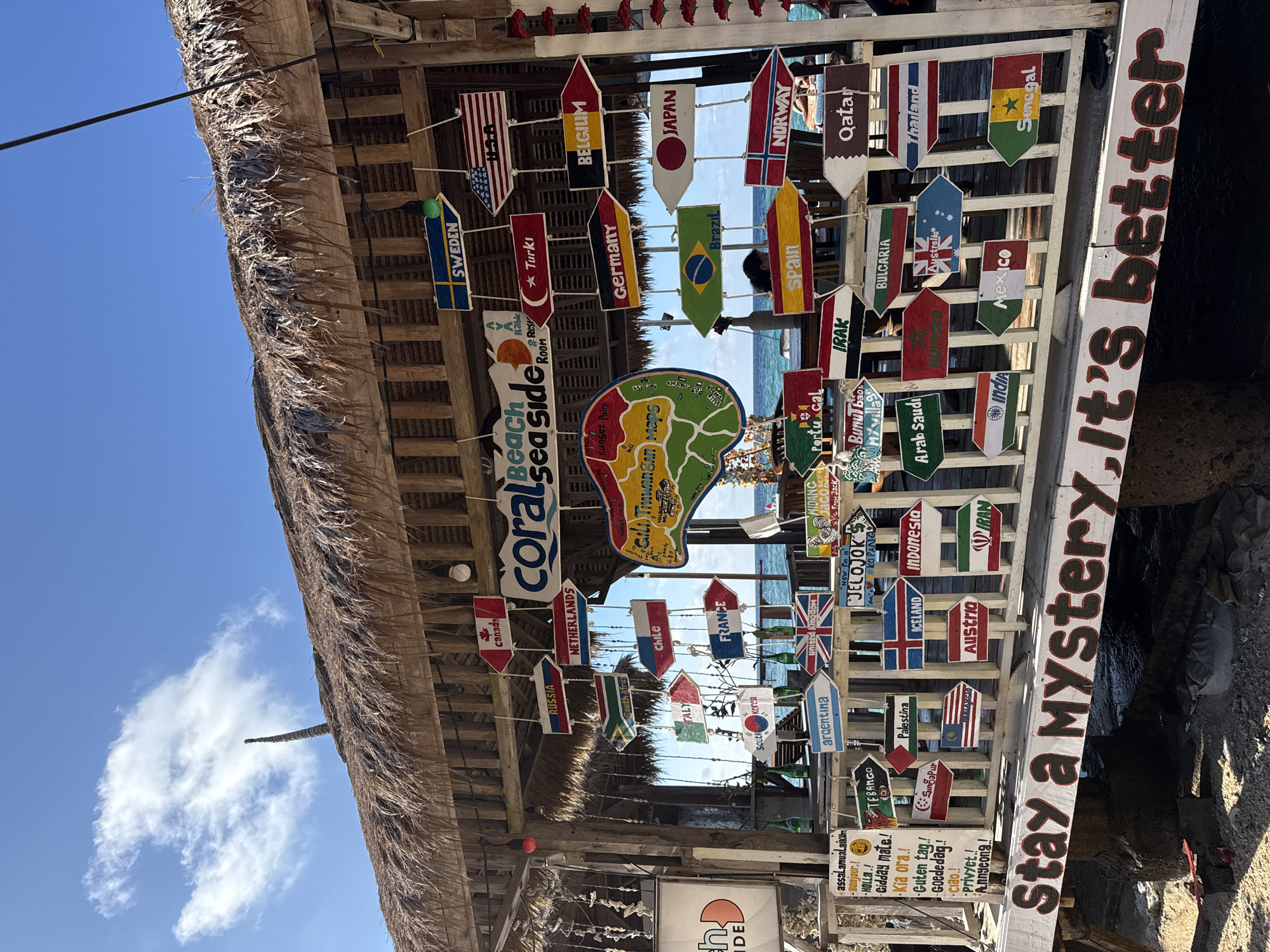 The image depicts a beachside structure, likely a bar or restaurant, at "coral beach sea side" in the Unknown city of Indonesia, as indicated by a sign displaying "Gili Trawangan Maps." The structure is covered with a thatched roof. Numerous arrow-shaped signs, each painted with the flag of a different country, are suspended from the structure. These signs include, but are not limited to, the flags of the USA, Belgium, Japan, Norway, Qatar, Thailand, Senegal, Australia, Mexico, India, Canada, Netherlands, Chile, France, the United Kingdom, Argentina, Palestine, Iceland, Austria, Iran, Germany, Brazil, Bulgaria, South Korea, and South Africa. A sign reads "Stay a Mystery, it's better."