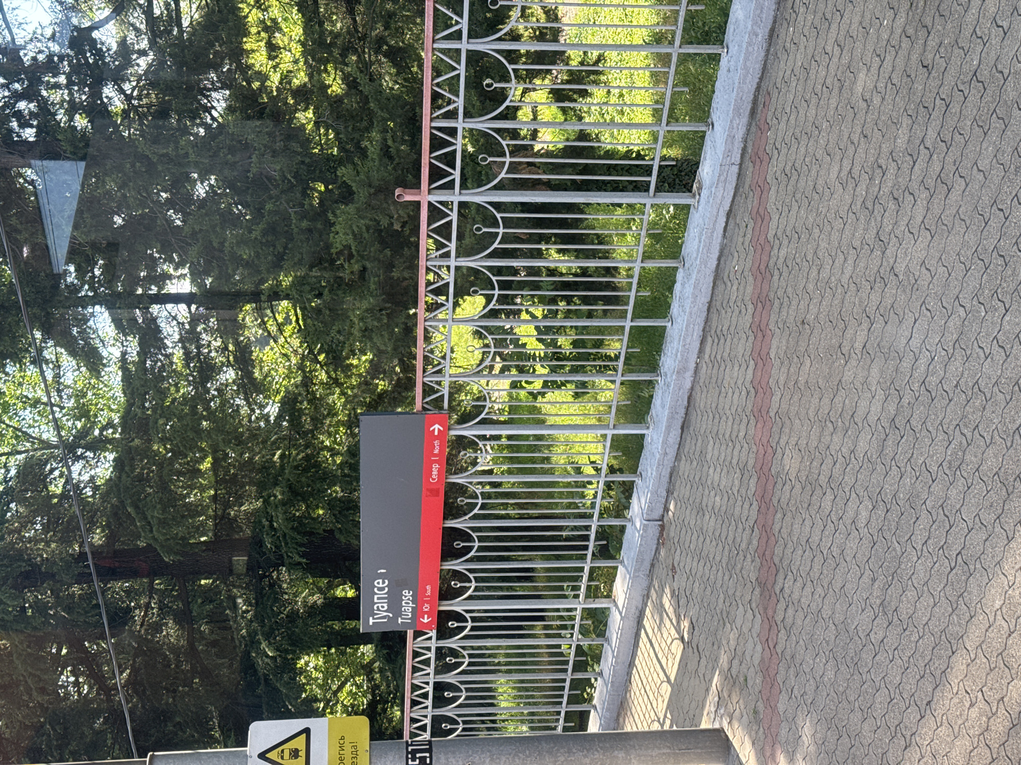 This image captures a sunlit outdoor scene at what appears to be a train station platform in Kholodnyi Rodnik, Russia. A prominent black sign with white and red lettering is central to the frame. The sign displays "Туапсе" (Tuapse) in large white Cyrillic script, with "Tuapse" in smaller English transliteration directly below it. A red bar beneath the station name indicates directions: an arrow pointing left with "Юг | South" (South) and an arrow pointing right with "Север | North" (North).
In the foreground, a gray and light-pink ornate metal fence runs horizontally, separating the paved platform area from a patch of green grass and dense evergreen trees. The fence features vertical bars and a top rail with repeating arched and circular decorative elements. The top rail of the fence is a faded pinkish-red color. The paved ground consists of interlocking grey pavers, interrupted by a distinct horizontal stripe of reddish pavers.
To the far left, a grey metal pole supports a yellow triangular warning sign depicting a black train icon. Partially visible text on this sign appears to be in Russian, likely a warning message. Behind the fence, a thick forest of tall, dark green coniferous trees stands under a bright sky, casting some shadows. There are no people visible in the scene, and no specific actions are taking place. The overall impression is one of a functional transit area surrounded by natural greenery on a clear day.
