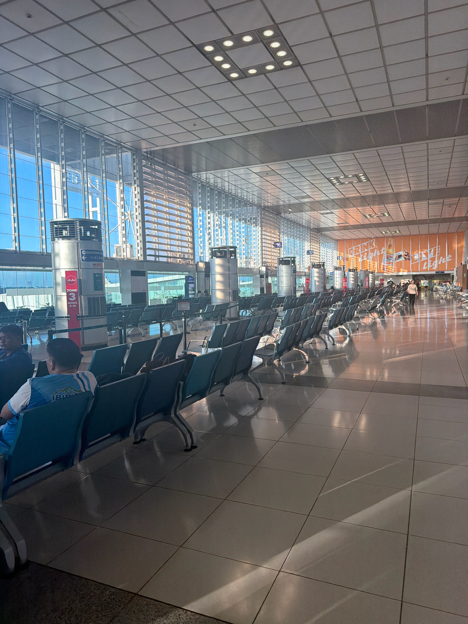 An interior view of a modern airport terminal waiting area in Pasay, Philippines. Rows of unoccupied, blue-seated chairs with metal frames are neatly arranged on a tiled floor. Sunlight streams through large glass windows, casting long shadows across the floor and creating reflections on the polished surface. In the background, several tall cylindrical structures with signage are visible, along with advertising posters on an orange wall. A few people are seated in the chairs, and one person is walking in the distance. The ceiling features a grid of square panels and recessed lighting. The overall atmosphere is one of quiet anticipation, typical of an airport departure lounge.