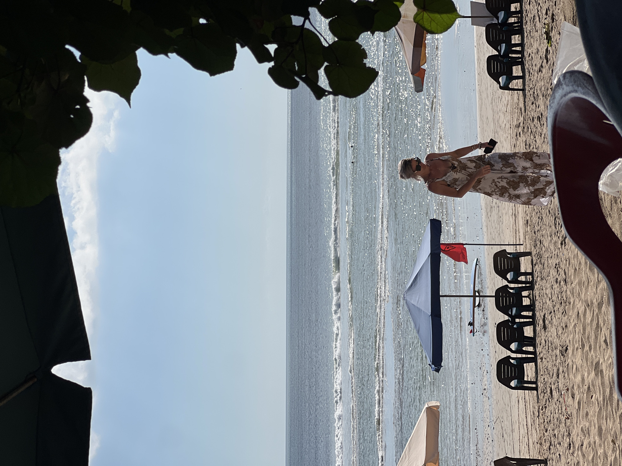 The image shows a beach scene in Kuta, Indonesia. In the foreground, a person wearing a long, patterned dress and sunglasses is walking on the sand, holding a phone. Behind them, several beach chairs are arranged under a large blue umbrella. A surfboard and a red flag are visible near the umbrella. Further in the background, the ocean stretches to the horizon under a clear blue sky with scattered clouds. Another umbrella is partially visible on the right side of the image, and foliage from a tree frames the top right corner.