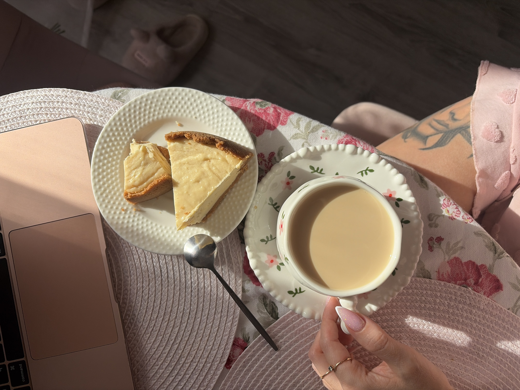 The image shows a laptop, a plate with two slices of cheesecake, a spoon, and a cup of coffee on a table. A woman is sitting next to the table and holding the cup of coffee with her right hand. She is wearing a pink outfit and has a tattoo on her right arm. The background is blurred, but it seems to be a room with a slipper in the top left corner.
It is not possible to determine if the location is Saint Petersburg, Russia or not.