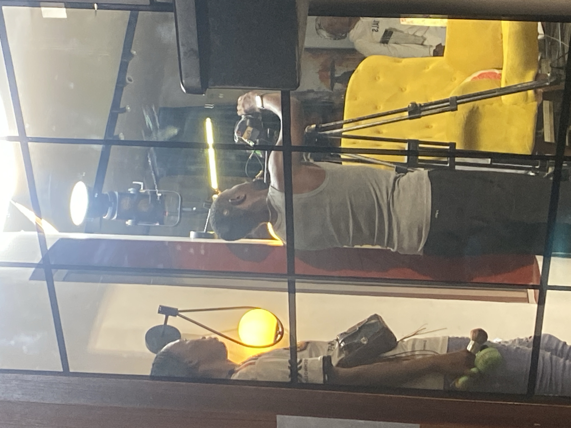 The image captures an indoor scene, divided into two distinct levels or areas by a horizontal glass partition. The photo appears to be taken through this glass, resulting in some reflections and minor distortions.
In the upper section, a person, seemingly a man, is actively filming or operating a camera. He wears a light-colored tank top and dark pants, with a smartwatch on his left wrist. To his left, a bright, vertical yellow strip light and a circular studio light are visible. In the background, a large, tufted yellow armchair is prominent, behind which another person in a white long-sleeved shirt with mirrored text "2JAT" (likely "TAJ") can be seen. Various equipment stands are also present, suggesting a studio or production environment.
The lower section displays a person lying on their back on a light-colored surface, possibly a bed or display platform. They are wearing a light-colored top and striped pants, appearing to be resting or asleep. A unique lamp with a spherical, glowing yellow bulb is positioned near their head, casting a warm light. A dark, rectangular bag or briefcase rests on their chest, and a green plush object is near their feet. Text that might read "ICHHOFER" is partially visible on their clothing.
The overall scene suggests an observation through a barrier, possibly depicting an art installation, a performance, or a studio setup where one activity (filming) is occurring in the foreground while a static or resting scene is presented in the background. The artificial lighting indicates an indoor setting.
