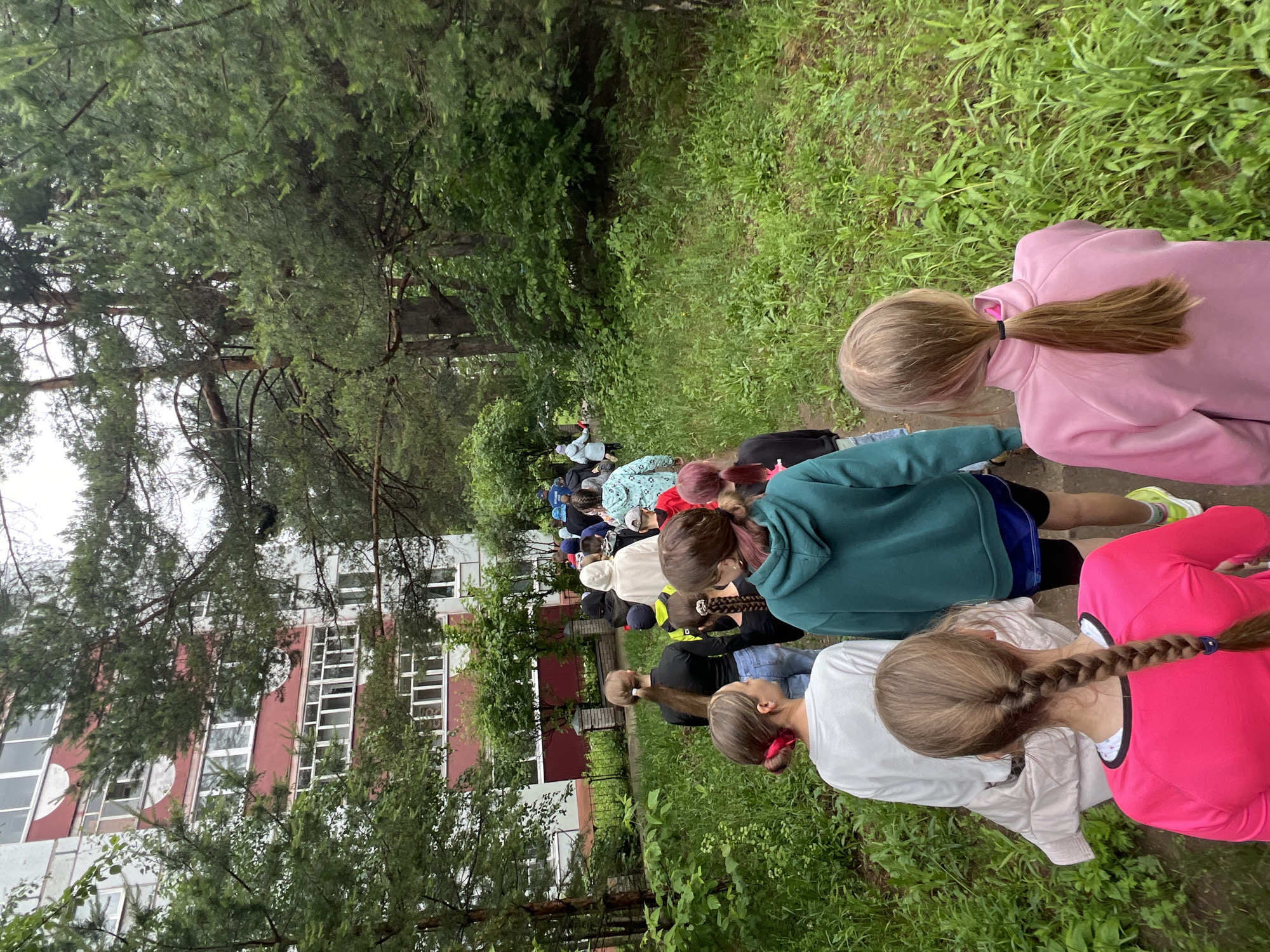 This media file depicts an outdoor scene featuring a line of children, predominantly girls, seen from behind. They appear to be walking or gathered on a narrow, unpaved path within a lush green environment. The children are dressed casually in hoodies and sweaters of various colors, including bright pink, teal, and white. Several have their hair tied back in ponytails or braids.
The path is bordered by dense vegetation, characterized by tall coniferous trees with needle-like leaves and thick green undergrowth, suggesting a forest, park, or natural setting. The lighting is diffused, indicative of an overcast day, and the vibrant greenery points to either spring or summer.
In the background, partially obscured by the trees, stands a large multi-story building. Its architecture includes distinct white vertical sections alternating with reddish-brown panels, and it features numerous rectangular windows. This structure could be a school, dormitory, or residential complex. The scene suggests an organized outdoor activity, such as a nature walk or group excursion, taking place in Chaikovsky, Russia. There is no visible text in the image.
