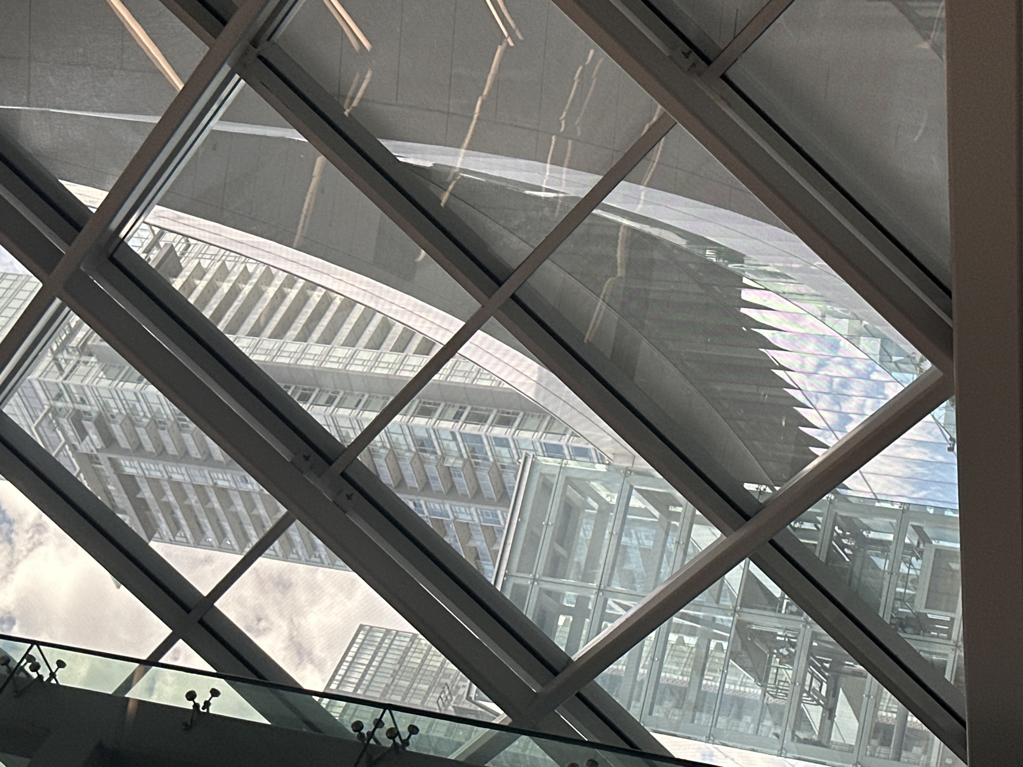 This image, captured inside the Surrey Central SkyTrain station in Burnaby, Canada, presents a view through a diamond-patterned glass ceiling looking upwards. The foreground is dominated by the angled white metal framing of the ceiling structure and the clear glass panes. Reflected in the glass and visible through it are several distinct architectural elements: to the left, a modern glass elevator shaft is visible, with multiple levels and visible elevator cars. Further into the background, a tall, multi-story residential building with numerous balconies and a glass facade is prominent. To the right, a large, curved, grey-toned building surface reflects light and what appear to be fluorescent light fixtures, suggesting an indoor commercial or public space. The sky, with clouds, is visible through some of the glass panels, indicating an open-air or semi-open-air structure. No people are clearly discernible in the image.