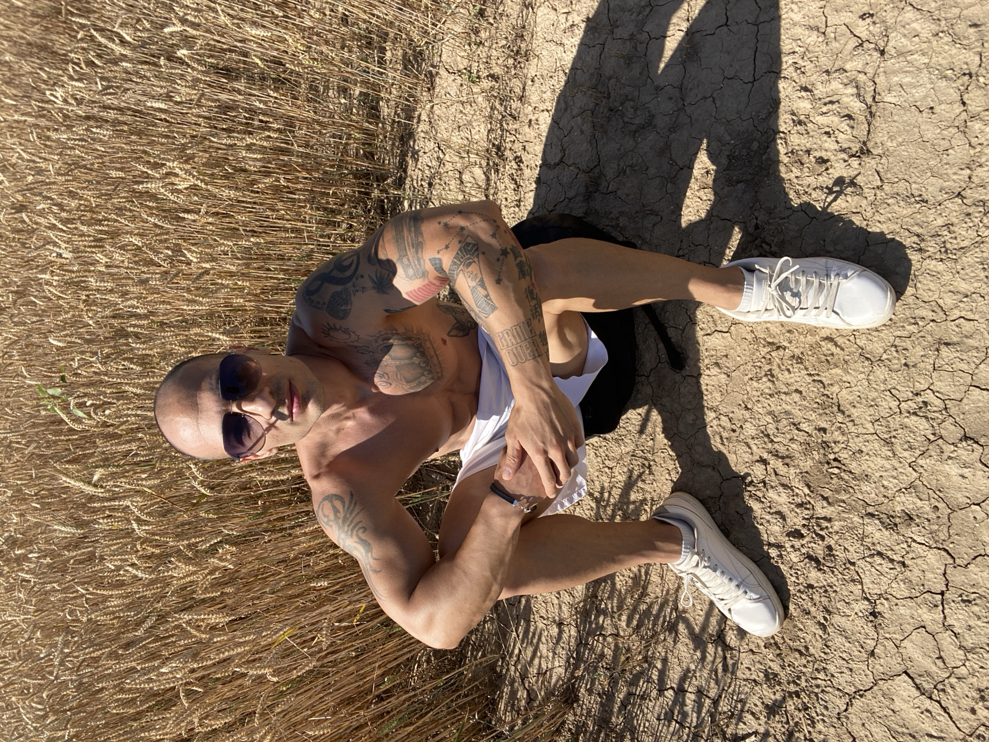 A bald man wearing sunglasses and white shorts kneels in a wheat field in Pleven, Bulgaria. He has tattoos on his arms and chest. The ground is dry and cracked. The sun is shining brightly, casting shadows.