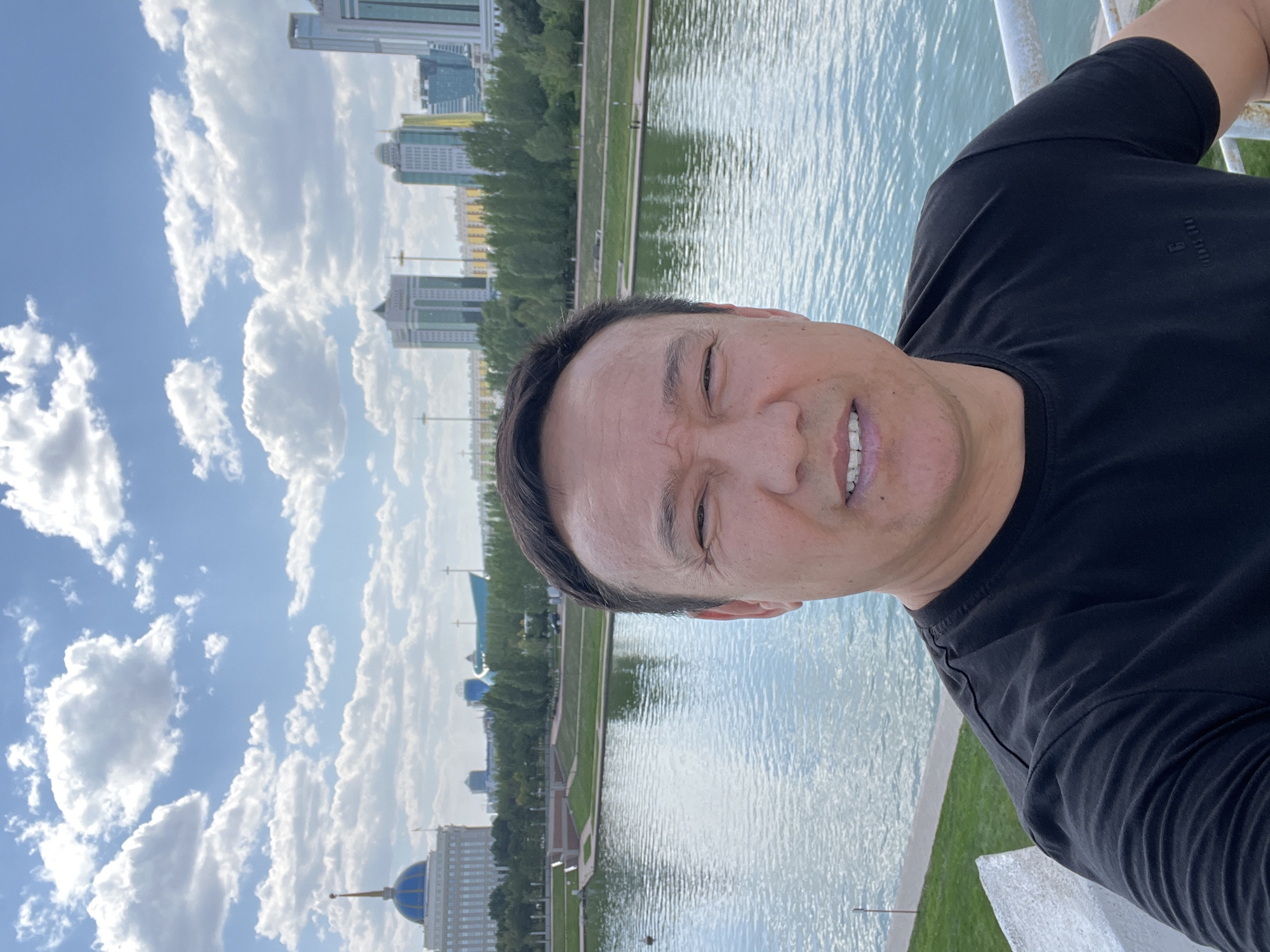 A man is taking a selfie outdoors in Astana, Kazakhstan. He is wearing a black t-shirt and is smiling slightly, with his teeth showing. Behind him, a river flows, with a park area and modern buildings visible in the distance. The sky is blue with scattered white clouds. The buildings include a prominent white structure with a blue dome and spire, resembling a presidential palace, and several tall, modern high-rise buildings. The scene appears to be a scenic urban landscape on a clear day.