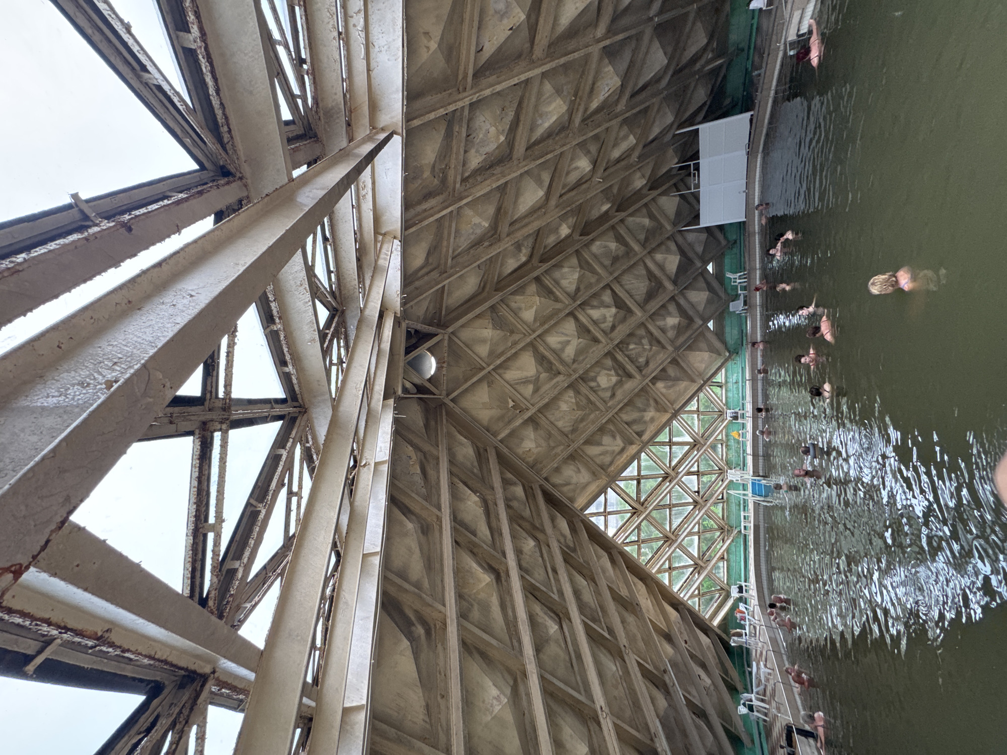 The image depicts an indoor swimming pool with a unique architectural design. The pool is filled with murky green water and several people are visible swimming or relaxing in it. The surrounding structure features a prominent, aged metal framework supporting a geometric, diamond-patterned ceiling. This ceiling appears to be made of concrete or a similar material, showing signs of wear and tear. Large windows or skylights are integrated into the structure, allowing natural light to enter, though the overcast sky suggests a cloudy day. The overall impression of the setting is one of an older, perhaps somewhat neglected, but functional recreational facility. There is no visible text in the image. The scene suggests a daytime activity, possibly a leisure outing or a visit to a thermal bath. The mood appears relaxed, with individuals enjoying the water. The setting is likely an indoor public pool, given the presence of multiple people and the architectural style.