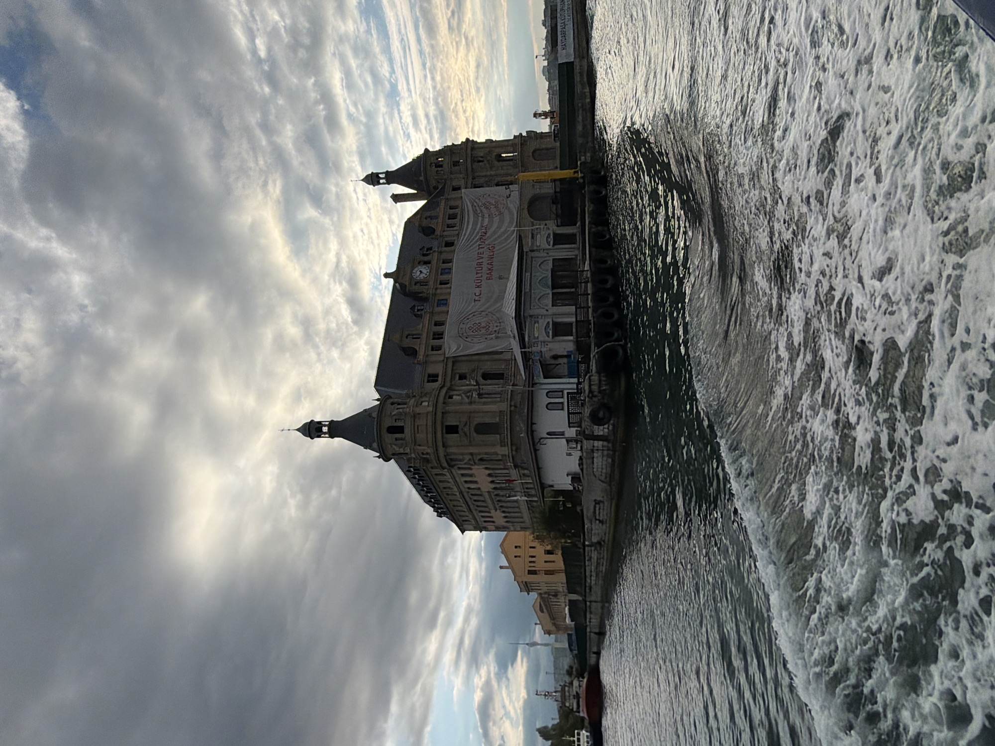 The image shows the historic Haydarpaşa Train Station in Kadıköy, Istanbul, Turkey. The station, a prominent landmark, is situated on the edge of the Bosphorus Strait. The architecture is grand, with classical European influences, featuring multiple towers, arches, and a prominent clock on its facade. A large banner hangs from the building, bearing Turkish text and symbols, likely related to a cultural or tourism event. The station is surrounded by water, and a wake is visible in the foreground, suggesting the photograph was taken from a moving boat. In the background, other buildings and a distant radio tower can be seen under a dramatic, cloudy sky. The overall scene depicts a waterfront view of an iconic piece of Istanbul's urban and historical landscape.