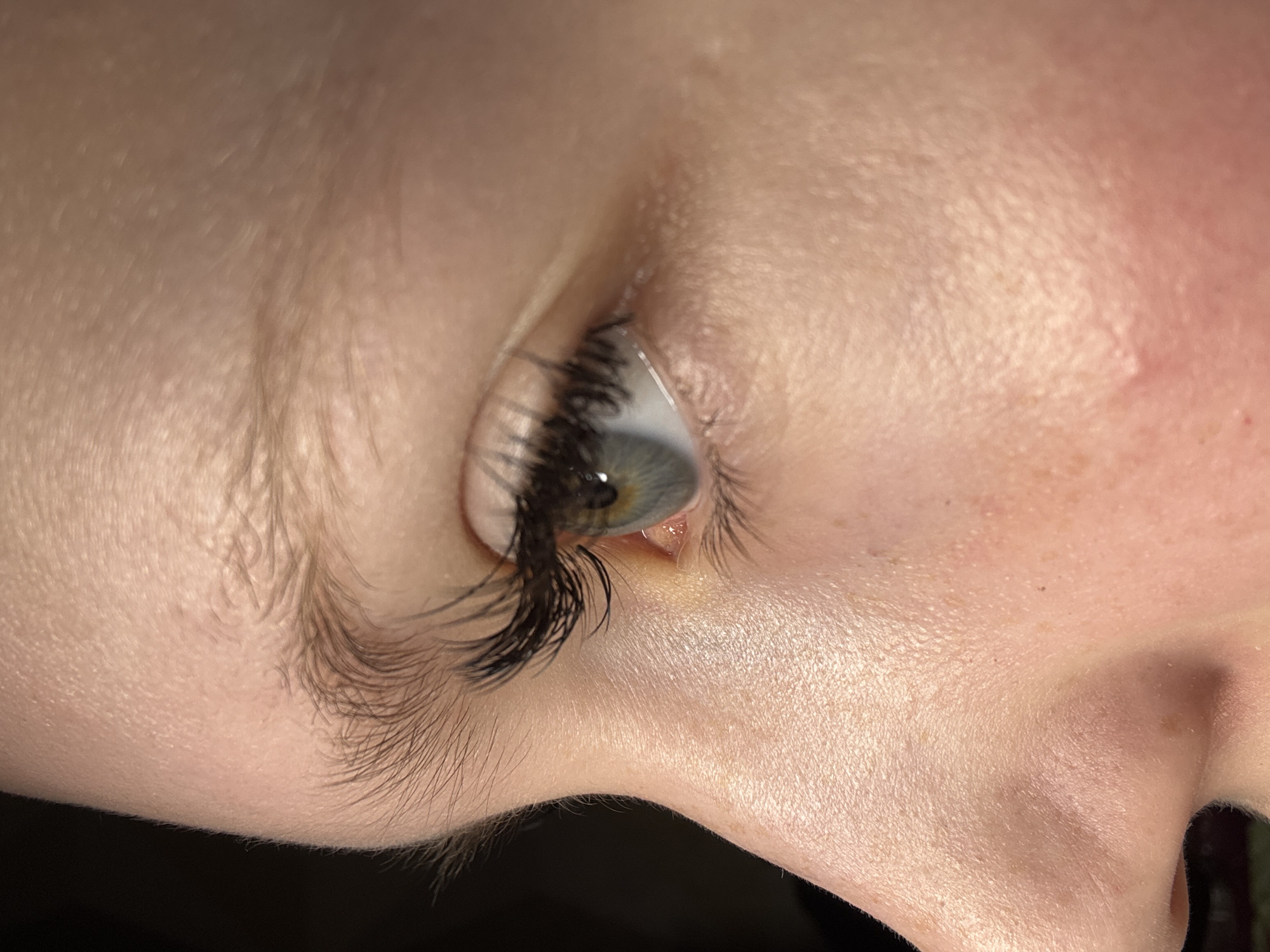 A close-up, angled view of a person's right eye and the surrounding facial area. The eye has a light iris with a dark limbal ring. Long, dark eyelashes are present, potentially enhanced with extensions. The skin tone is fair and shows fine wrinkles and pores. Light brown hair is visible along the eyebrow and temple region. The image does not reveal any location information related to the city of Chaikovsky, Russia, or any other specific environment.