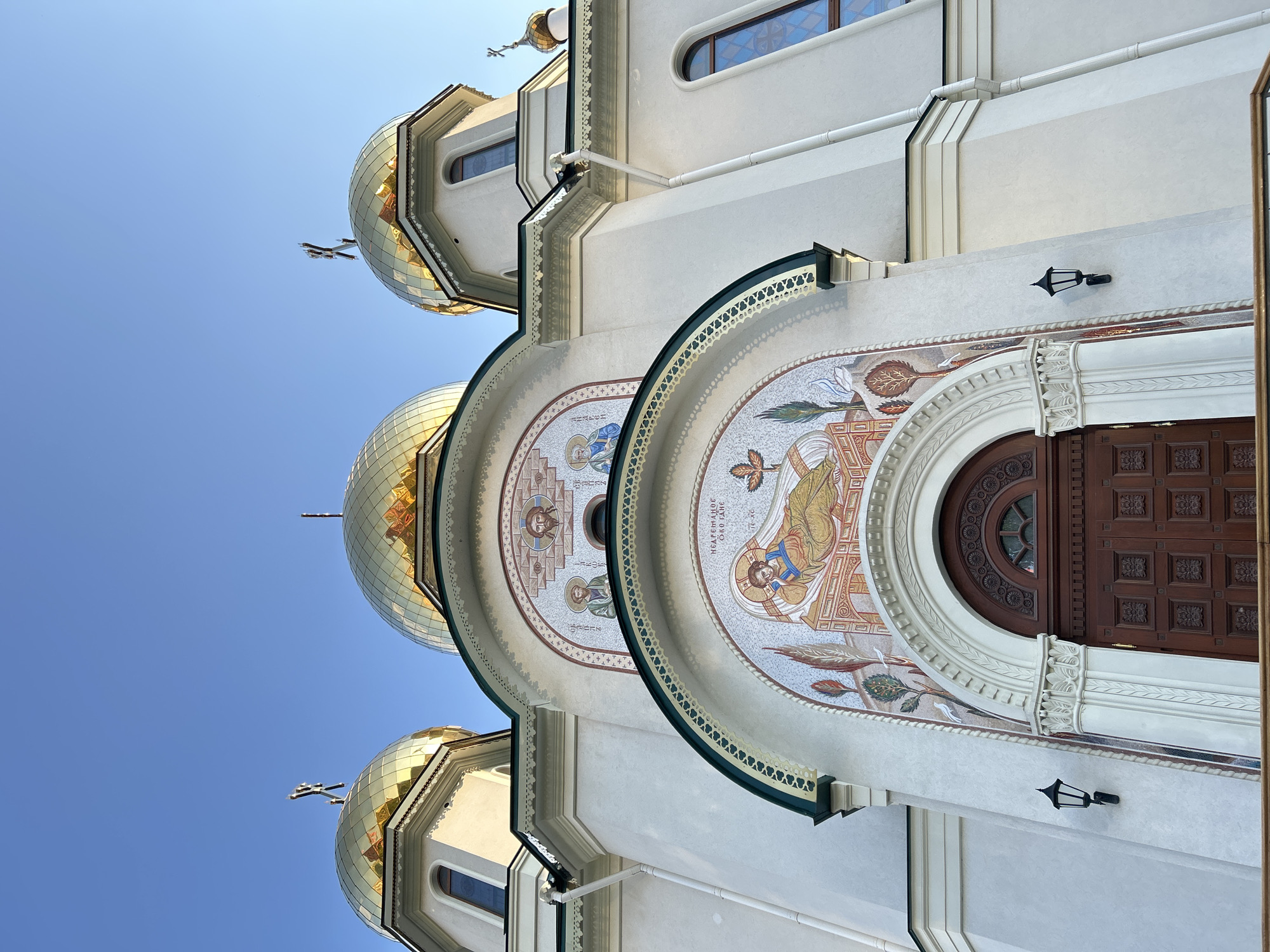 The image depicts the exterior of an Orthodox church, likely located in Saint Petersburg, Russia, given the architectural style and golden onion domes. The building is white with intricate detailing, including mosaic-style artwork above the arched doorways. One doorway is visible, constructed with dark wood. Lampposts are affixed to the exterior wall. The sky is clear and blue.