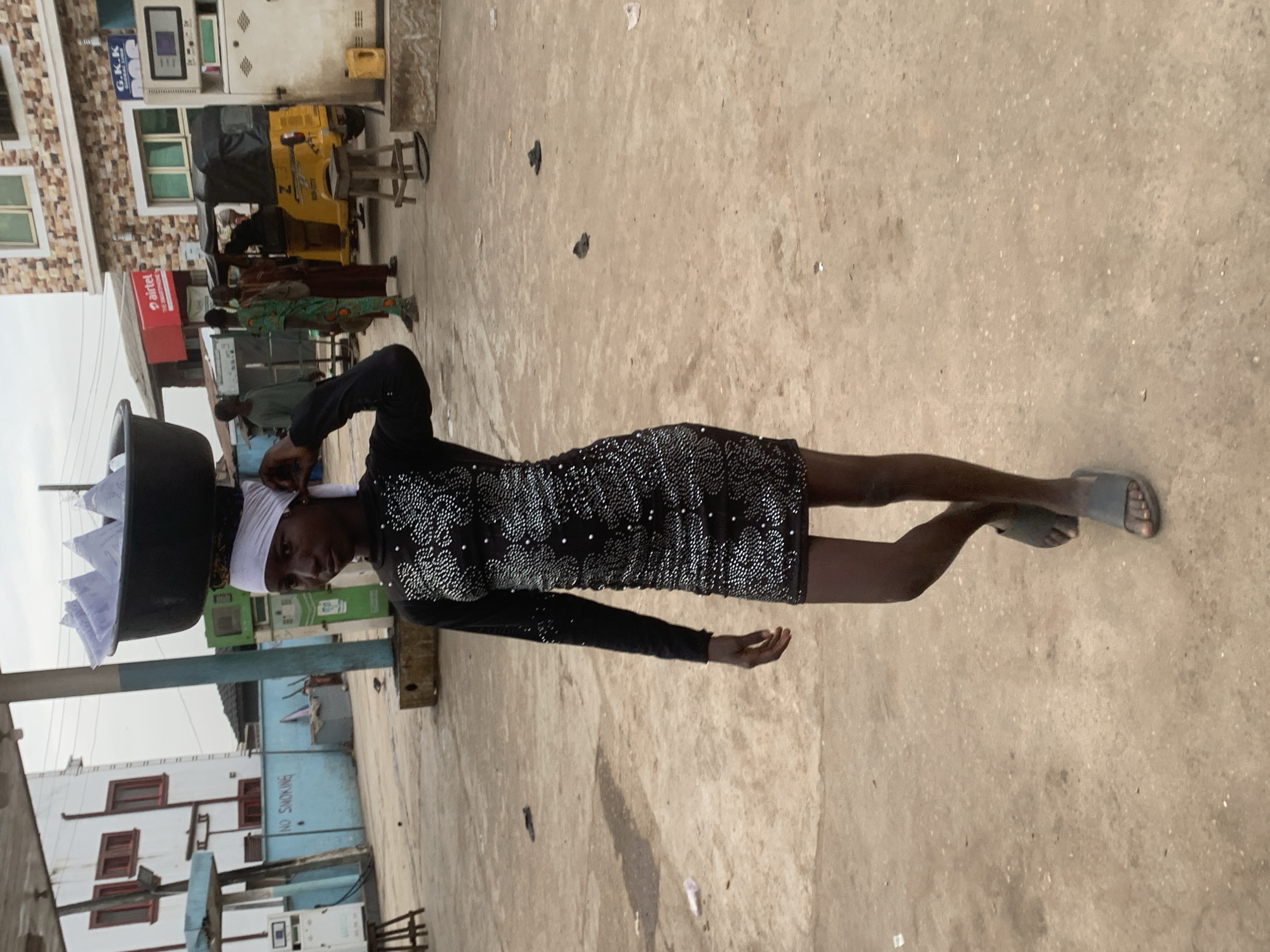 The image shows a young person standing on a dusty, outdoor ground. They are wearing a black, long-sleeved dress adorned with silver embellishments and a white headwrap. The person is carrying a large black basin on their head, filled with white items. To their left, a yellow three-wheeled vehicle is parked in front of a building with stone cladding and windows. Several other people, some in traditional attire, are visible in the background near the vehicle. Behind them, buildings with various architectural styles and colors are present, including one with a sign that appears to read "Airtel". Another building in the distance has blue walls and a sign with red lettering that says "NO SMOKING". The scene suggests a bustling environment, possibly a market or a street in a town. The lighting indicates it could be daytime, with diffused light suggesting an overcast sky. The overall impression is a candid moment capturing daily life in Shagamu, Nigeria.