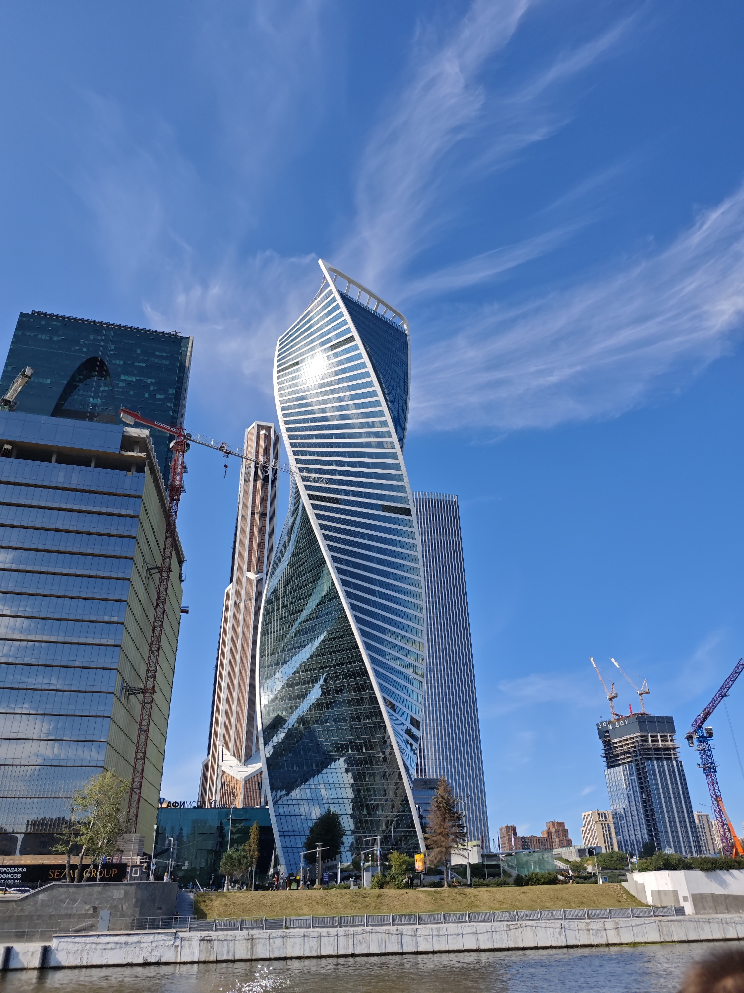 This image depicts the Moscow International Business Center, also known as Moscow City, a commercial district in western Moscow, Russia. The scene is dominated by several modern skyscrapers, including the distinctive twisting building of the Neva Towers. To the left, a partially constructed glass skyscraper is visible, with cranes indicating ongoing development. The sky is a clear blue with wispy white clouds. In the foreground, a body of water, likely the Moskva River, reflects the sunlight and the surrounding architecture. The riverbank is lined with a concrete embankment and a grassy slope. Several trees are sparsely distributed along the embankment. The image captures a sunny day in a rapidly developing urban landscape.