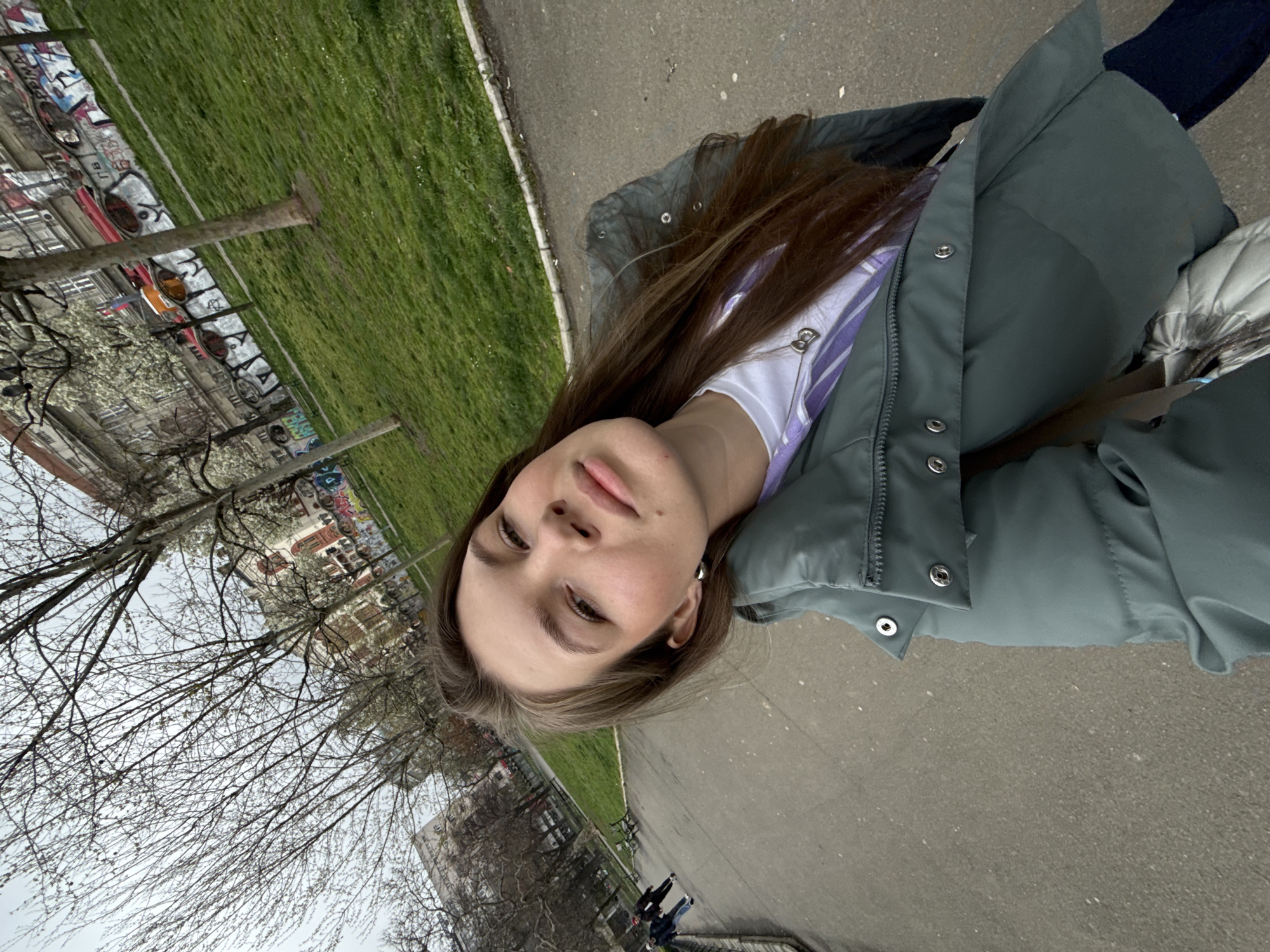A person is seen from a low angle on an urban path, likely a selfie. The individual has long brown hair and is wearing a green puffer jacket over a white and purple striped shirt. Behind the person is a grassy area bordered by a road and buildings, and trees with budding spring foliage. There are apartment buildings in the background with graffiti. The location is the city of Belgrade, Serbia. Other individuals can be seen in the distance on the path.