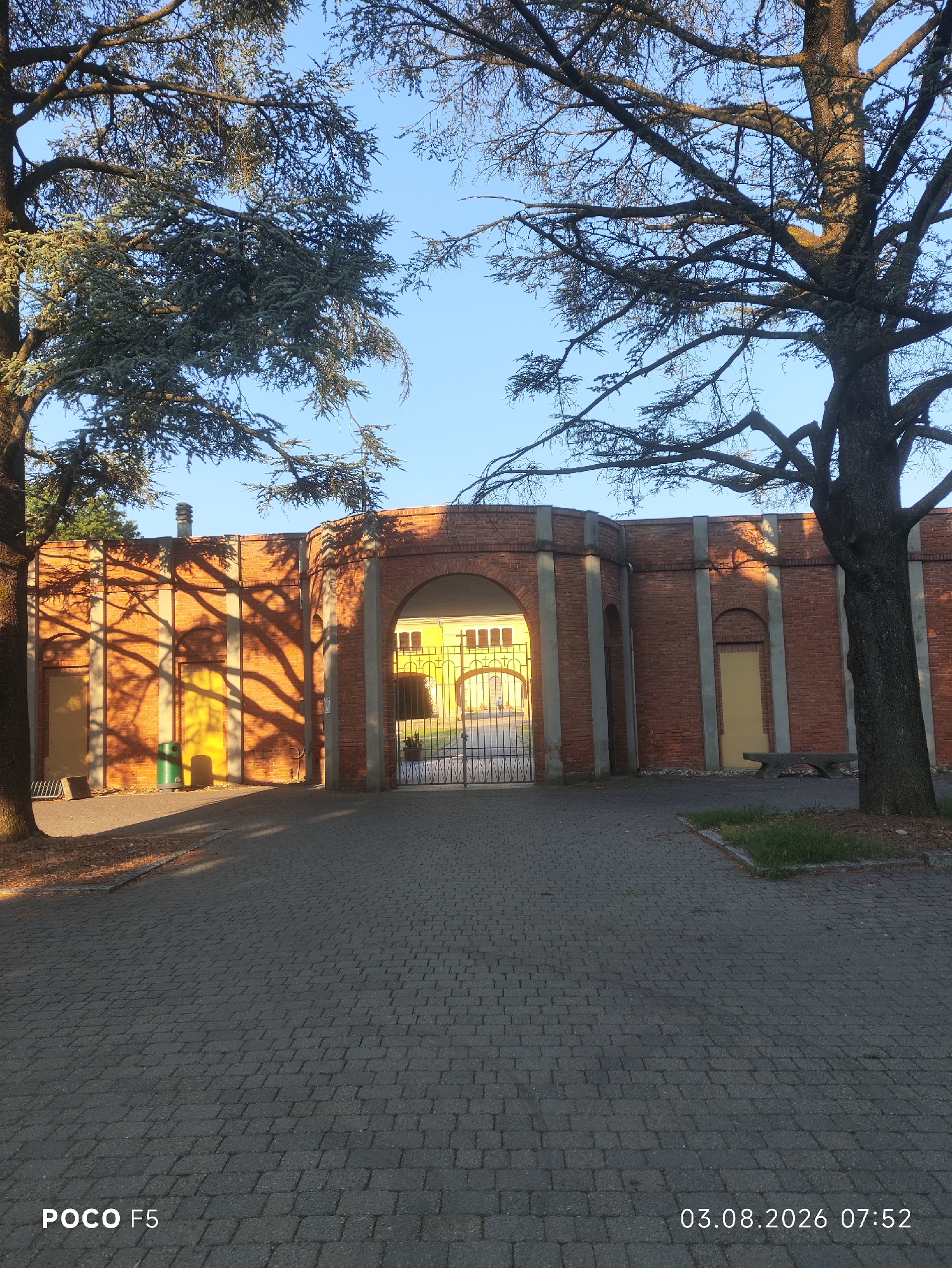 The image depicts a view of a brick wall with an arched gateway in Collecchio, Italy. Two large trees frame the scene, casting long shadows across the paved ground and the wall. The wall has several pilasters and arched recesses, with some doors visible. Through the main archway, a yellow building and a metal gate are discernible. A green trash can and a bench are present near the wall. The sky is clear and blue, indicating daytime. The timestamp on the bottom right corner reads "03.08.2026 07:52" and the text "POCO F5" is on the bottom left.
