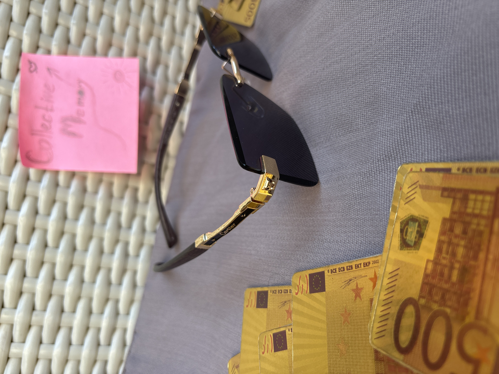 A close-up shot depicts a still life arrangement. A pink sticky note with the inscription "Collective Memory" and a sun drawing sits on the left side. A pair of Cartier sunglasses is positioned centrally, with the brand name visible on the temple. Several gold-colored 500 Euro decorative notes are stacked on the right. The objects rest on a light gray fabric surface. The background consists of a woven white pattern. This arrangement is indoors, likely in a cafe or restaurant. The image does not show a city or location in Budva, Montenegro.