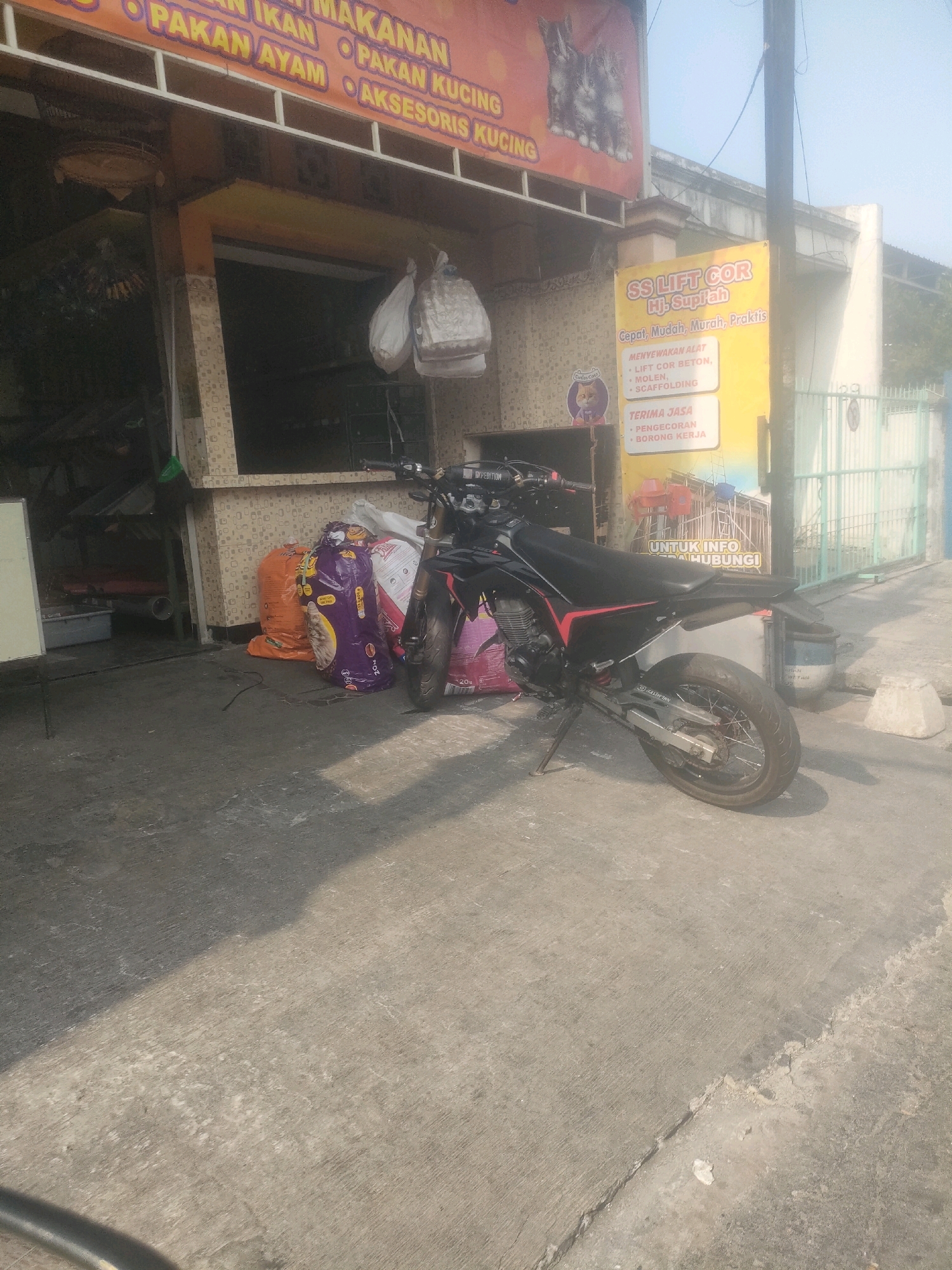 The image depicts a street scene in Surabaya, Indonesia, in front of a store with an orange awning. The awning displays text in Indonesian, including "MAKANAN IKAN," "PAKAN AYAM," "MAKANAN," "PAKAN KUCING," and "AKSESORIS KUCING," indicating it sells pet food and accessories. To the right of the store, a yellow banner advertises "SS LIFT COR Hj. Supriyah," offering services like renting lifting equipment ("LIFT COR BETON," "MOLEN," "SCAFFOLDING") and general contracting ("PENGECORAN," "BORONG KERJA"). Below this, it states "UNTUK INFO HUBUNGI" (for information, contact). A black and red dirt bike is parked on the concrete sidewalk in front of the store, leaning on its kickstand. Several large bags of pet food, one purple and one orange, are stacked near the bike. The environment appears to be a commercial area with a slightly hazy sky.