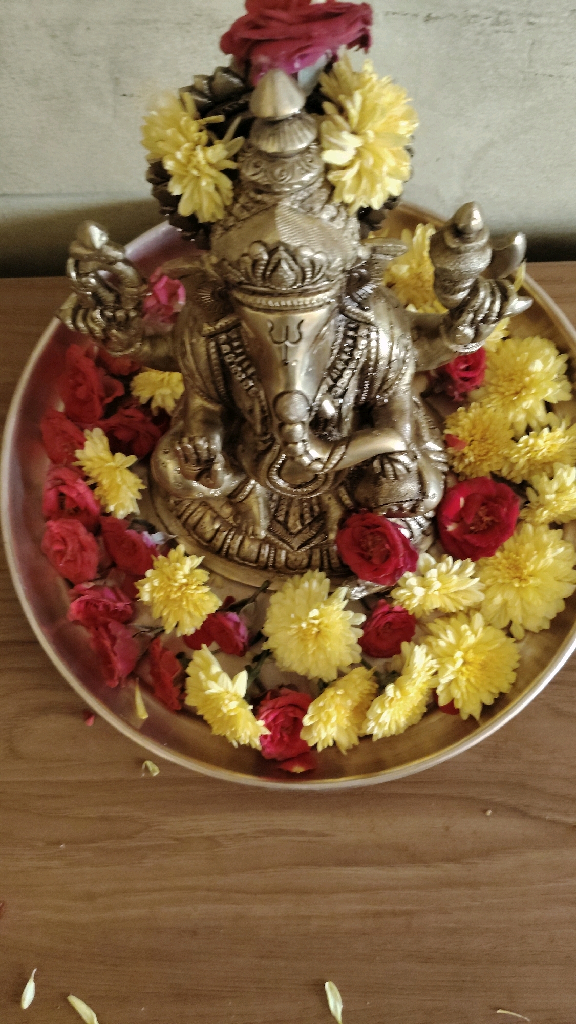 A gilded statue of the Hindu deity Ganesha is placed on a brass plate adorned with red roses and yellow chrysanthemums. The plate is situated on a wooden surface, likely a table. The statue depicts Ganesha in a seated posture, with his trunk curled and his hands holding various implements. The deity is decorated with floral garlands and a red cloth is draped over its head. The background is a textured, light-colored wall. There are no people or animals visible in the image. The scene appears to be set indoors, in a residential or devotional setting, possibly within a household in Bengaluru, India, given the cultural context of Ganesha worship. The lighting is soft and diffused, creating a warm and serene atmosphere.