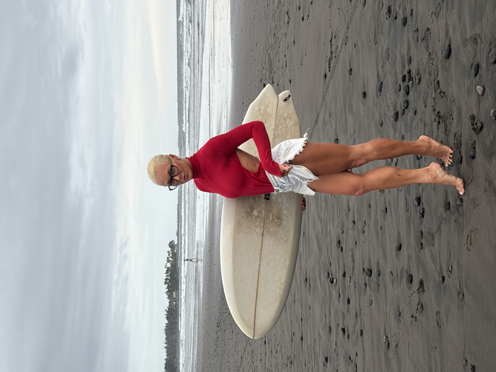 A woman with blonde hair and glasses is posing with a surfboard on a beach. She is wearing a red long-sleeved top and white shorts. The beach has dark sand and small rocks. The ocean is in the background, with waves visible. There is vegetation near the shoreline. The location appears to be a beach in an unknown city.