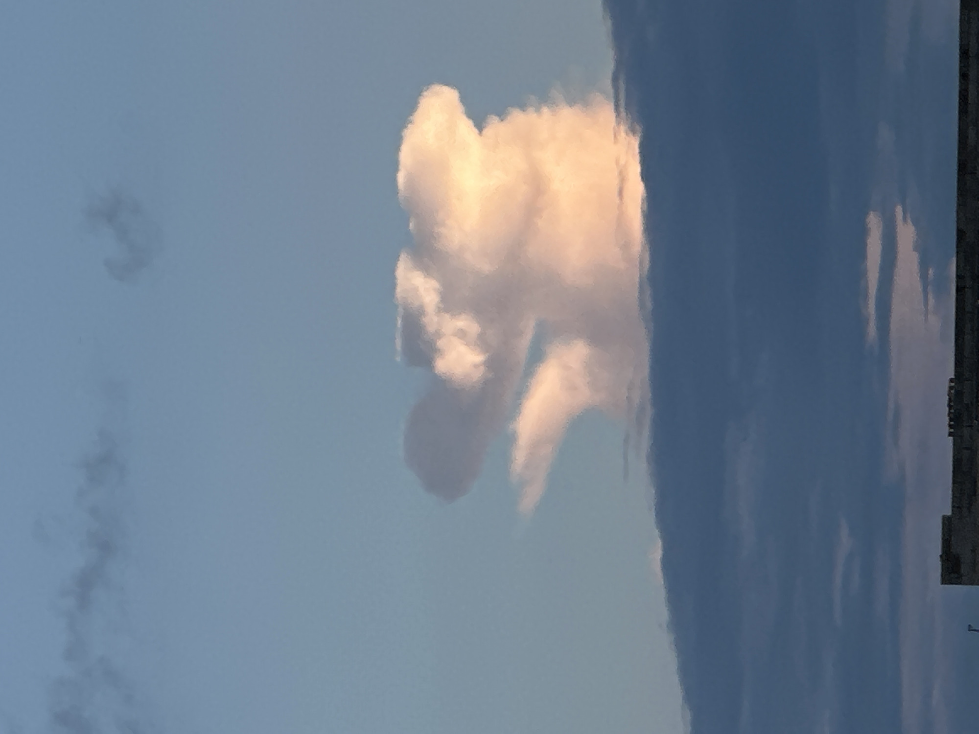 A vertical shot captures a cloud formation in the sky above Tiskre, Estonia. The cloud is lit from the side by the sun, giving it a peachy hue. It's shaped somewhat like a person. In the foreground, a layer of darker, lower clouds obscures the horizon, and at the very bottom of the frame, the top of a building is visible.