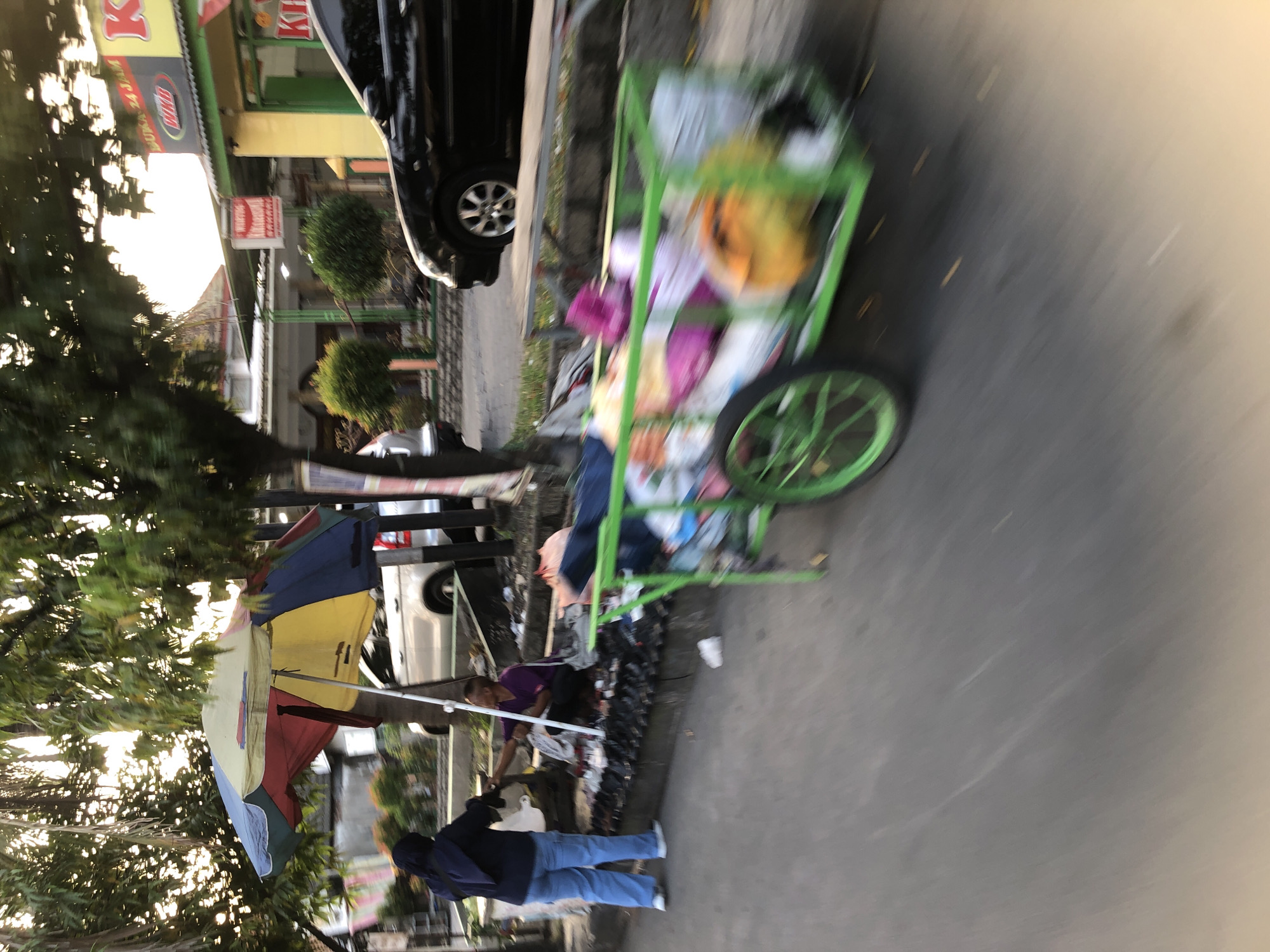 The image depicts a street scene in Tenggulunan, Indonesia, with motion blur indicating movement. In the foreground, a green cart overflowing with various items, including colorful fabrics and what appear to be household goods, is being pushed along the street. A black car is parked on the right side of the street, with its front tire visible. On the left side of the street, a woman wearing a dark blue hijab and light blue jeans stands near a colorful umbrella, interacting with a person sitting on the ground. This person, wearing a purple shirt, is surrounded by an assortment of items laid out on the ground, likely for sale. The background shows buildings, trees, and more vehicles, all slightly blurred due to the motion of the camera. The overall scene suggests a bustling street market or roadside vendors.