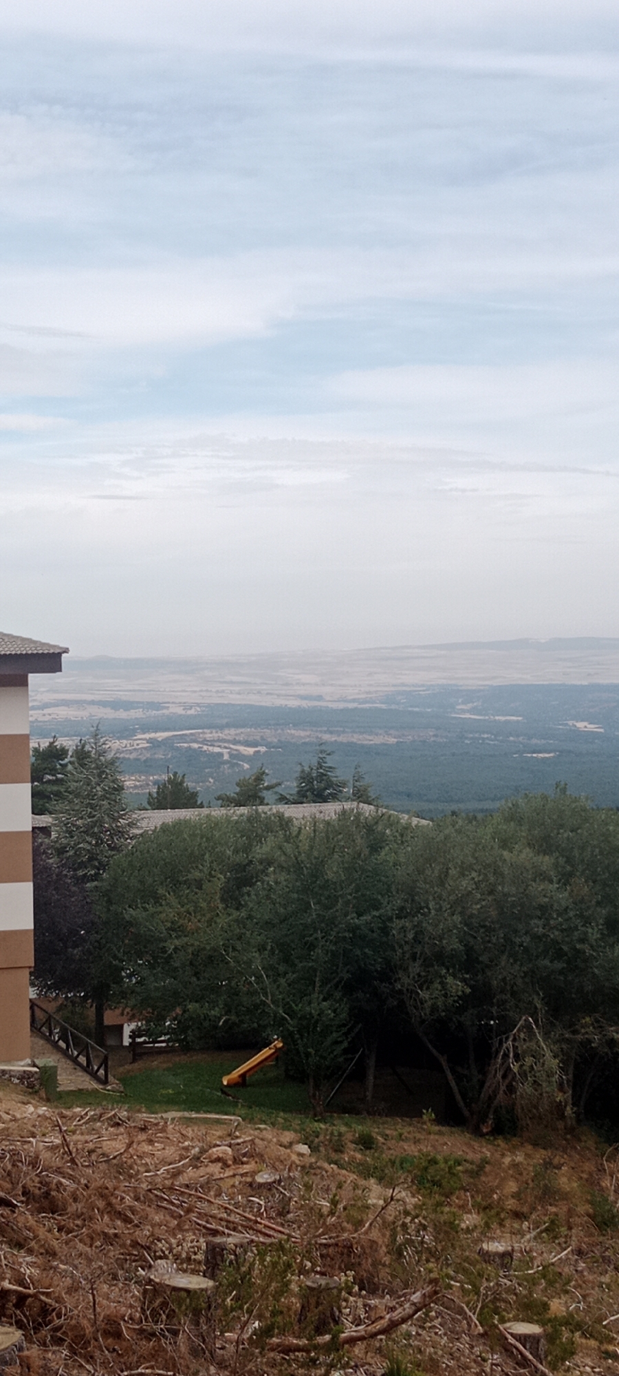 The image captures a scenic view from a residential building, likely located in Cerezo de Arriba, Spain. In the foreground, a sloped area is covered with uprooted trees, branches, and stumps, suggesting recent deforestation or land clearing. A portion of a building with horizontally striped brown and white siding is visible on the left, with a wooden fence and railings nearby.
The middle ground features a lush green lawn dotted with trees and a yellow slide. This area appears to be part of a garden or playground. Further back, a dense forest stretches across the landscape, leading to a distant, hazy vista of rolling hills and plains under a pale blue sky with scattered clouds. The overall impression is one of a rural or semi-rural setting with a contrast between cleared land and natural greenery. There are no people or animals visible in the image.