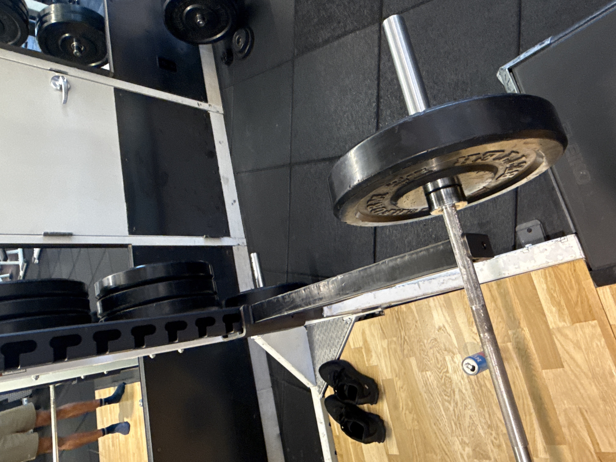 The image depicts a gym setting in West Hollywood, California. The scene includes weightlifting equipment: a barbell loaded with a single black weight plate, and a rack with a stack of additional weight plates. A pair of black shoes sits on the floor near the weight rack. Part of a person's legs and feet are visible, wearing socks and standing on the floor, partially obscured by the weight rack. The flooring consists of rubber mats and wooden planks. A beverage can is attached to the barbell with tape.