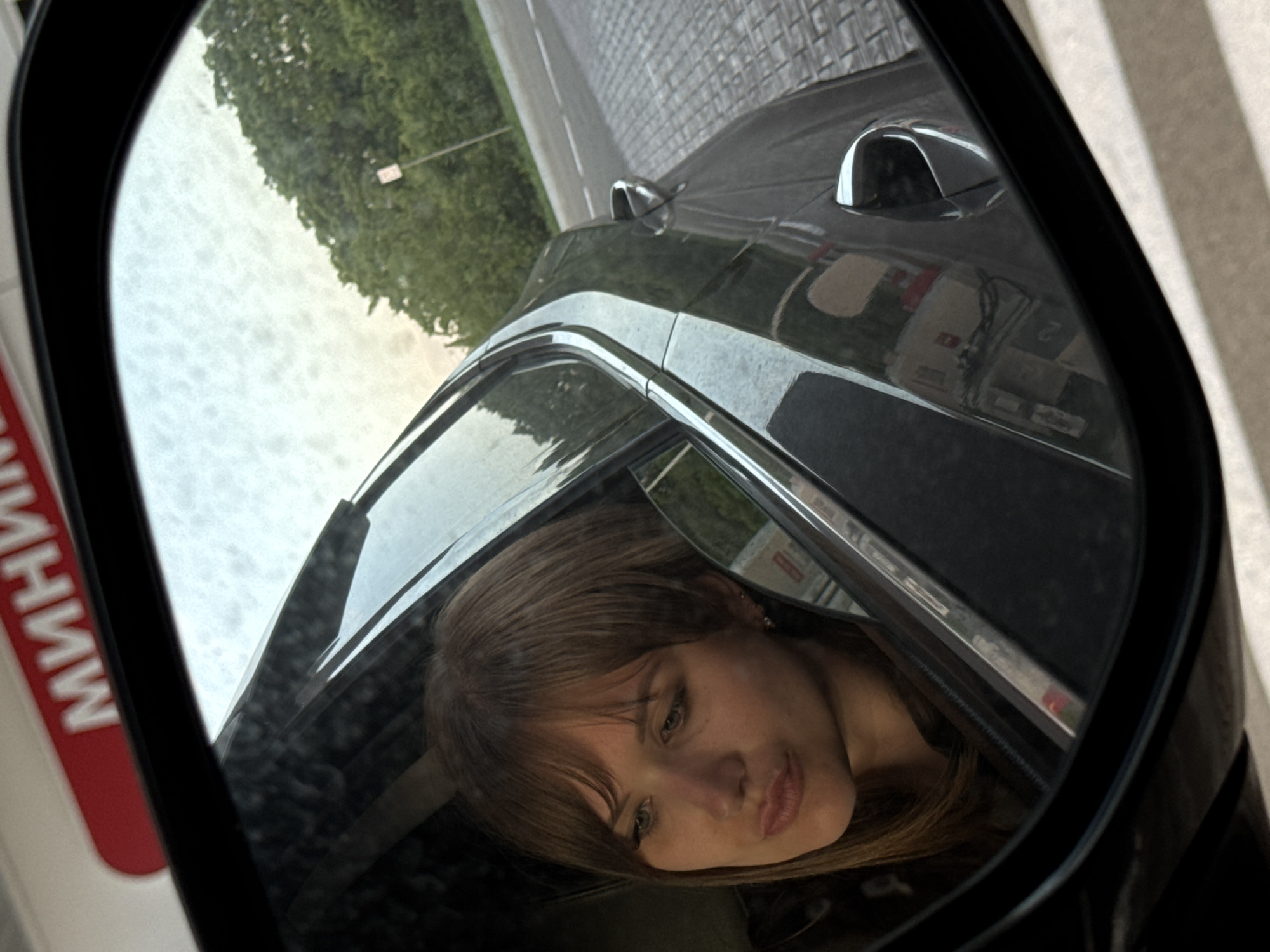 A side-view mirror of a dark-colored car reflects the face of a woman with shoulder-length brown hair and bangs. The car is parked near a street with trees visible in the distance. Reflected in the car's paint is a building with the word "МИНИМ" written in Cyrillic. The location is Vinogradny, Russia.