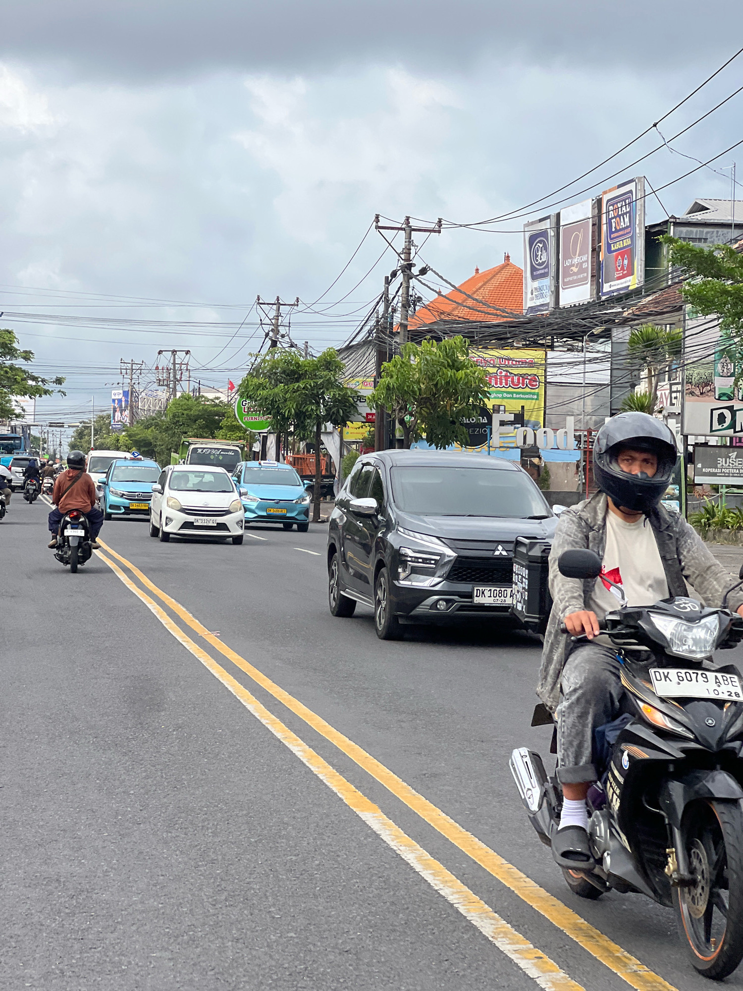 The image shows a street scene in Denpasar, Indonesia, with a line of vehicles on an overcast day. In the foreground, a man wearing a helmet, jacket, and jeans rides a motorcycle, facing right and looking towards the camera. His left leg is visible, wearing a white sock and black slide sandal. The motorcycle has a license plate reading "DK 6079 ABE". Further down the road, a dark grey SUV with a license plate "DK 1080 A" is also visible, traveling in the same direction. Several other vehicles, including blue cars and a white car, are in the traffic flow. The road has yellow double lines. On the sides of the road, there are buildings with signs, including one for "Royal Foam" and another with "Furniture". Power lines are strung across the top of the frame, supported by utility poles. Trees and vegetation are present along the roadside. The overall atmosphere appears to be a typical urban traffic scene.