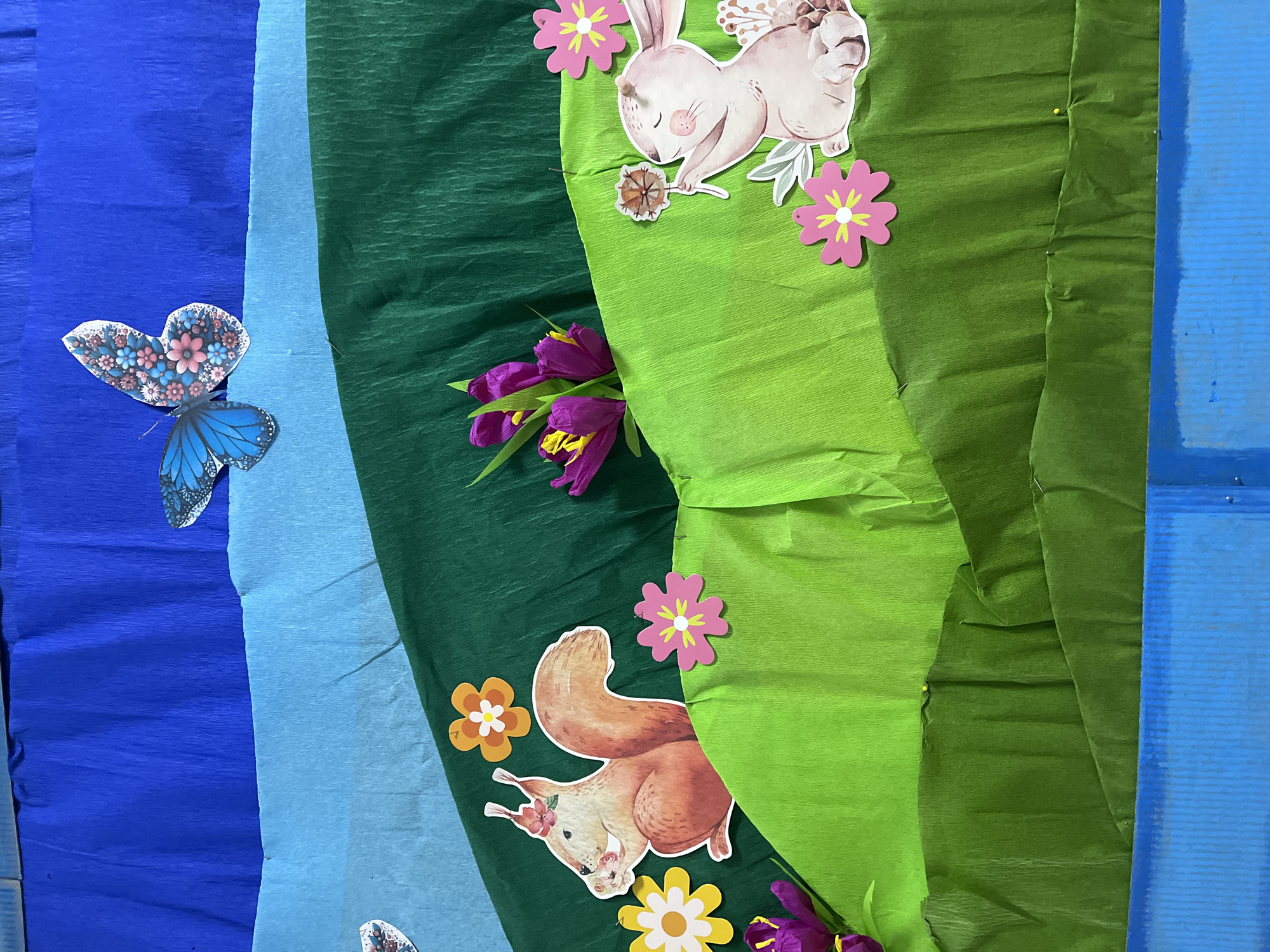 The image displays a vibrant scene with multiple layers of textured crepe paper in shades of blue and green forming a backdrop. A decorative butterfly with blue wings speckled with flower patterns rests on the blue crepe paper. In the green area, several printed images of animals, specifically a rabbit and a squirrel, are featured along with cut-out paper flowers of varying sizes and colors. Fresh purple tulips are also present, adding to the natural aesthetic of the scene. No specific location or city can be determined from this image.