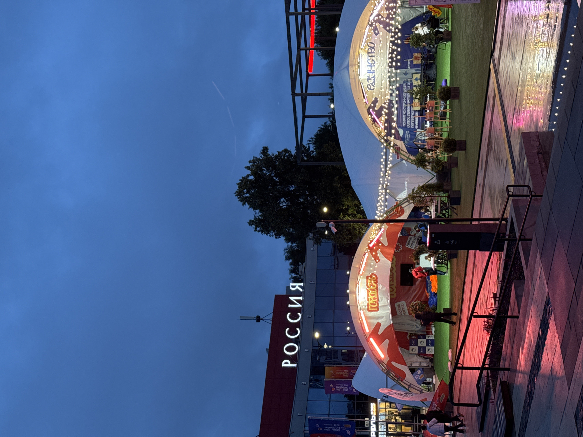 The image depicts an outdoor event space at dusk in Izhevsk, Russia. In the background, a building with a large sign spelling "РОССИЯ" (RUSSIA) in illuminated white letters is visible. The building has a dark facade with large windows. In the foreground, there are two large white tents with illuminated red and white curved structures on their fronts. The tent on the left has text in Russian that includes "гордость" (pride), and the tent on the right has text that includes "единство" (unity). Both tents are decorated with strings of fairy lights.
Under the tents, there are artificial grass areas with people mingling, some sitting at tables, and what appears to be an art display or exhibition in progress. Several potted plants are also present. In front of the tents, there is a paved walkway that is wet and reflects the red and pink lights from an unseen source, suggesting recent rain or intentional lighting. A dark metal railing and stairs are in the immediate foreground. A few individuals are walking in the scene, and some are standing or seated. The sky is a deep blue, indicative of twilight, with faint contrails from an aircraft visible.