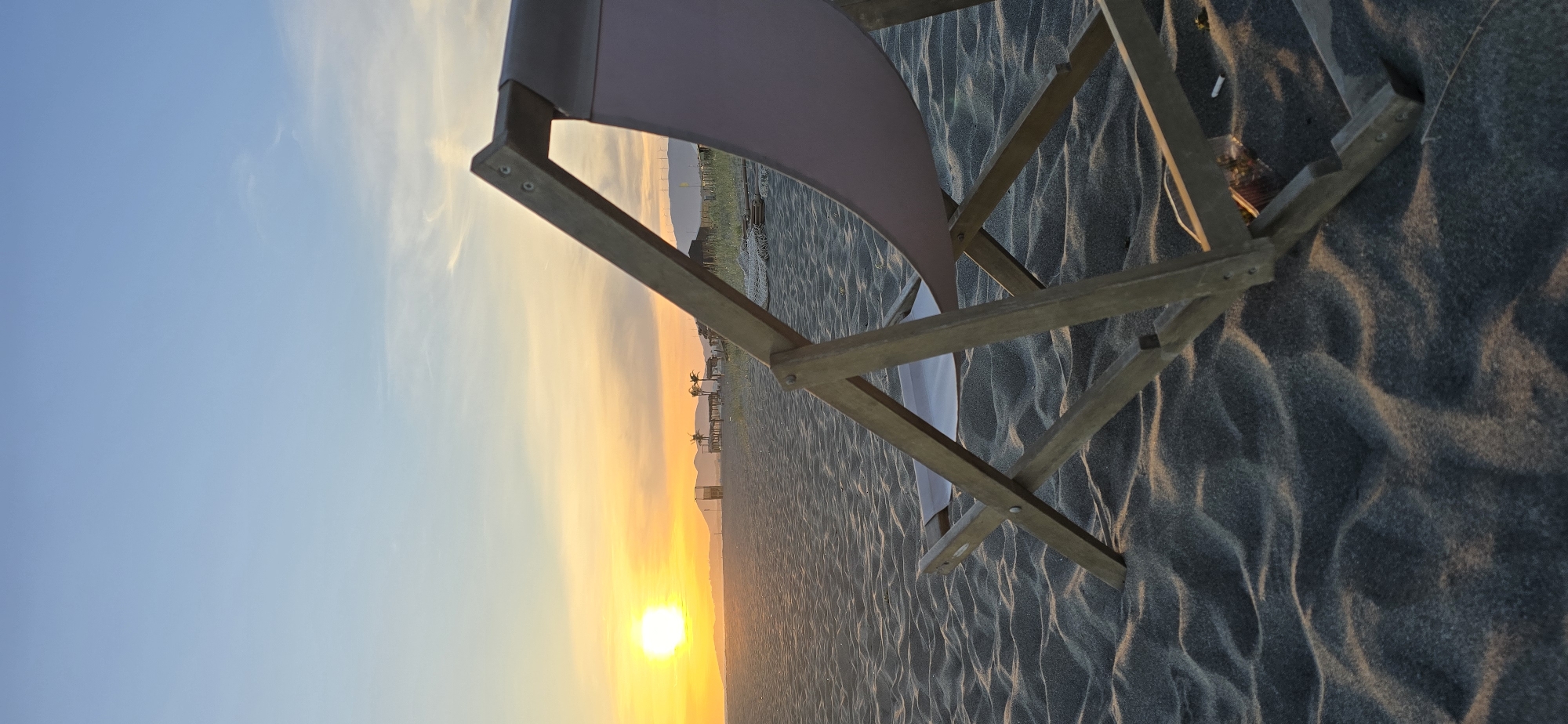 A landscape photo from Gornji Štoj - Shtoji i Epërm, Montenegro shows a beach chair on dark sand with a bright orange sun setting over the horizon. The sky is blue fading into yellow. The chair frame is made of light-colored wood, and its seat is a light pink fabric. The sand has ripples and dark shadows.