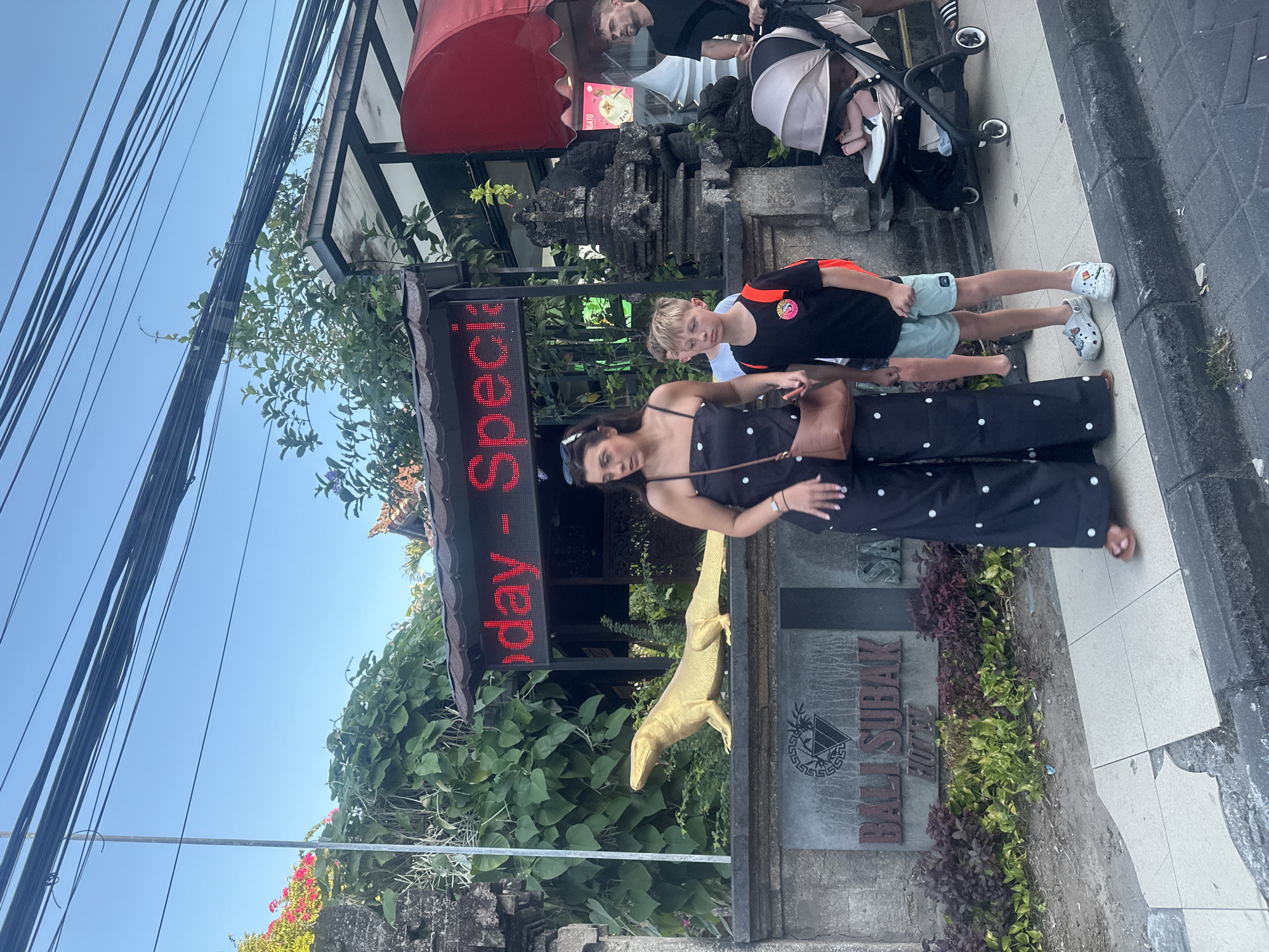 In Kuta, Indonesia, a group of people are gathered on a sidewalk. In the foreground, a woman in a black polka-dot jumpsuit and a brown bag is standing. Beside her, a young boy with blond hair, wearing a black t-shirt with orange accents and light blue shorts, is also standing. Behind them, a baby in a stroller is visible with an adult partially obscured. Further back, another adult is seen behind a stone structure.
The scene appears to be a street, with a building entrance behind the people. A large, illuminated red sign displays text vertically. A golden statue of a lizard-like creature stands prominently in front of the building. The sky is clear and blue, suggesting a daytime setting. The weather seems to be sunny and warm.
The sign displays the text "Today - Special" in red LED letters. Part of a building sign in the background reads "BALI SUBAK HOTEL". The overall impression is a lively street scene in a tourist area, possibly depicting people on vacation or out for the day.
