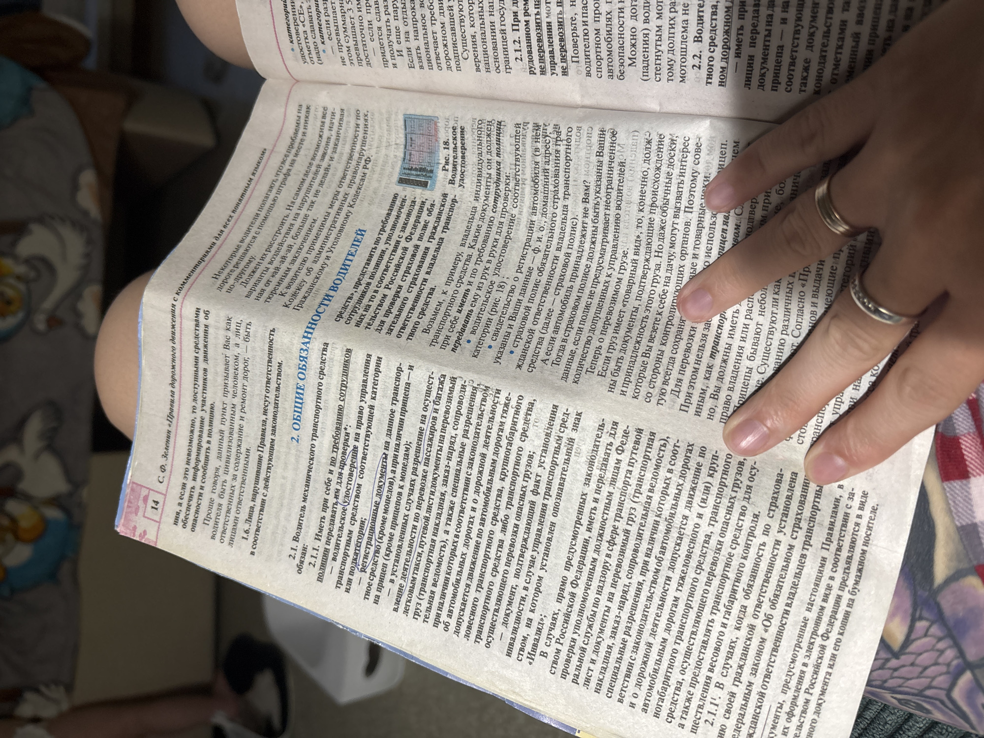 The image shows a close-up of a book with Russian text, held open by two hands. The book appears to be a manual or guide related to driving regulations, as indicated by headings like "Правила дорожного движения" (Traffic Rules) and "ОБЩИЕ ОБЯЗАННОСТИ ВОДИТЕЛЕЙ" (General Duties of Drivers). The text discusses vehicle documents, driver responsibilities, and legal consequences. The lighting suggests an indoor setting, possibly a room with overhead light. The background is out of focus, but appears to be a surface like a bed or couch, with a portion of a cartoon character visible. The image does not provide any information related to the city of Naberezhnye Chelny, Russia.