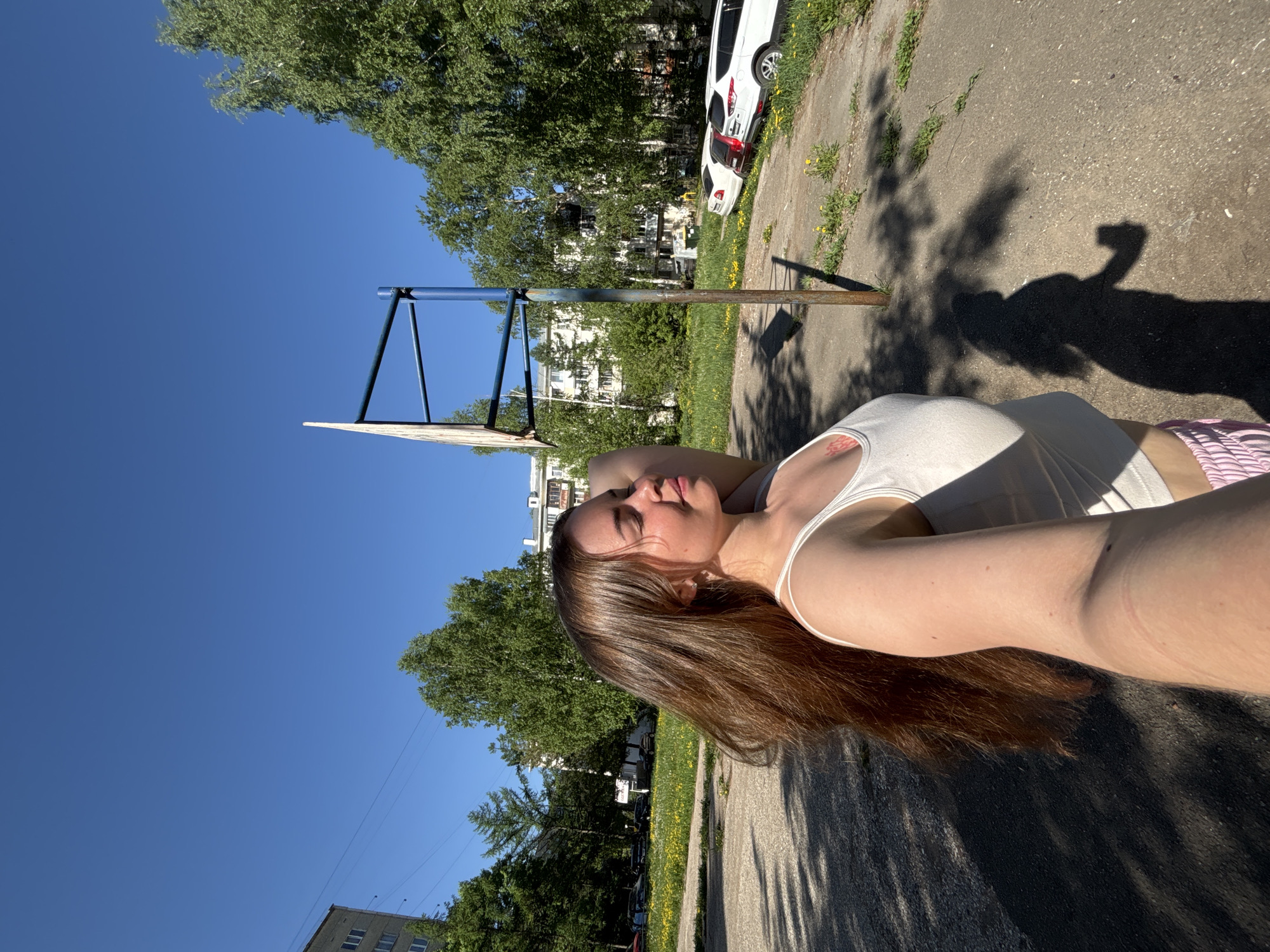 A woman is lying on a concrete surface next to a basketball court under a sunny sky. The basketball hoop is visible on the left side of the image. Behind the hoop, there are green trees and buildings. In the background, there is a white car. A shadow is cast on the ground. The location is Izhevsk, Russia.