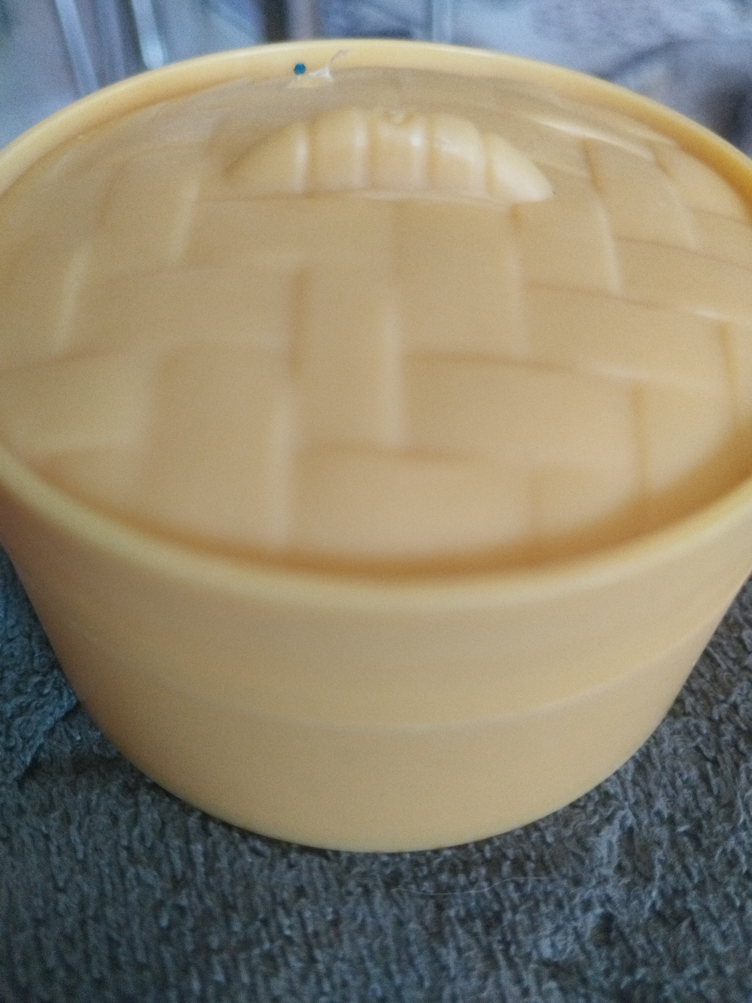 The image shows a close-up of a light yellow, plastic lidded container. The lid has a woven basket-like texture molded into its surface. A small blue speck is visible on the lid near the top left edge. The container is resting on a textured, dark grey surface, possibly a blanket or fabric. The background is blurred, showing hints of blue and grey. There is no indication of the location in Oskemen, Kazakhstan. The image is a still photograph with no discernible actions or interactions.
