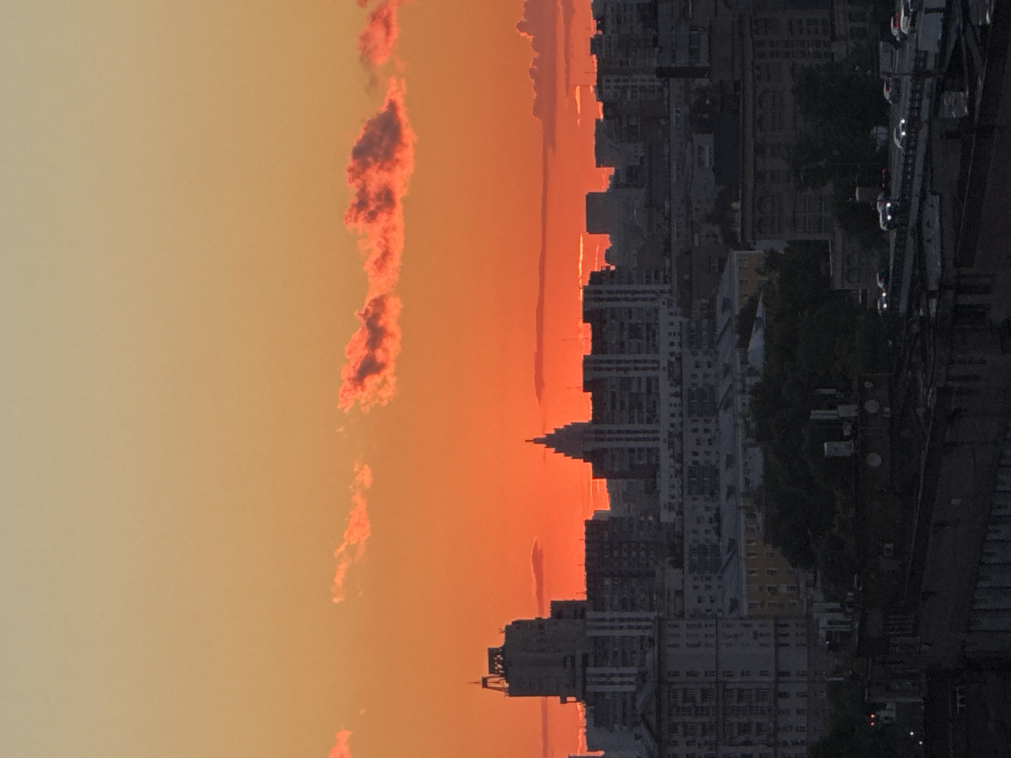 This is a vertical, high-angle shot of a cityscape at sunset. The sky is filled with warm orange and yellow hues, with streaks of pink clouds. The right side of the frame is dominated by the silhouette of a dense urban landscape with numerous tall buildings. The architecture appears to be a mix of modern high-rises and older apartment blocks. Trees are visible between some of the buildings, adding a touch of green to the scene. In the foreground, to the far right, a street or elevated roadway is lined with cars, their headlights and taillights visible as red and white dots, suggesting traffic. The overall impression is one of an active city at the end of the day, bathed in the dramatic light of the setting sun. The scene is likely in Moscow, Russia, given the building styles and the presence of a dense urban environment.