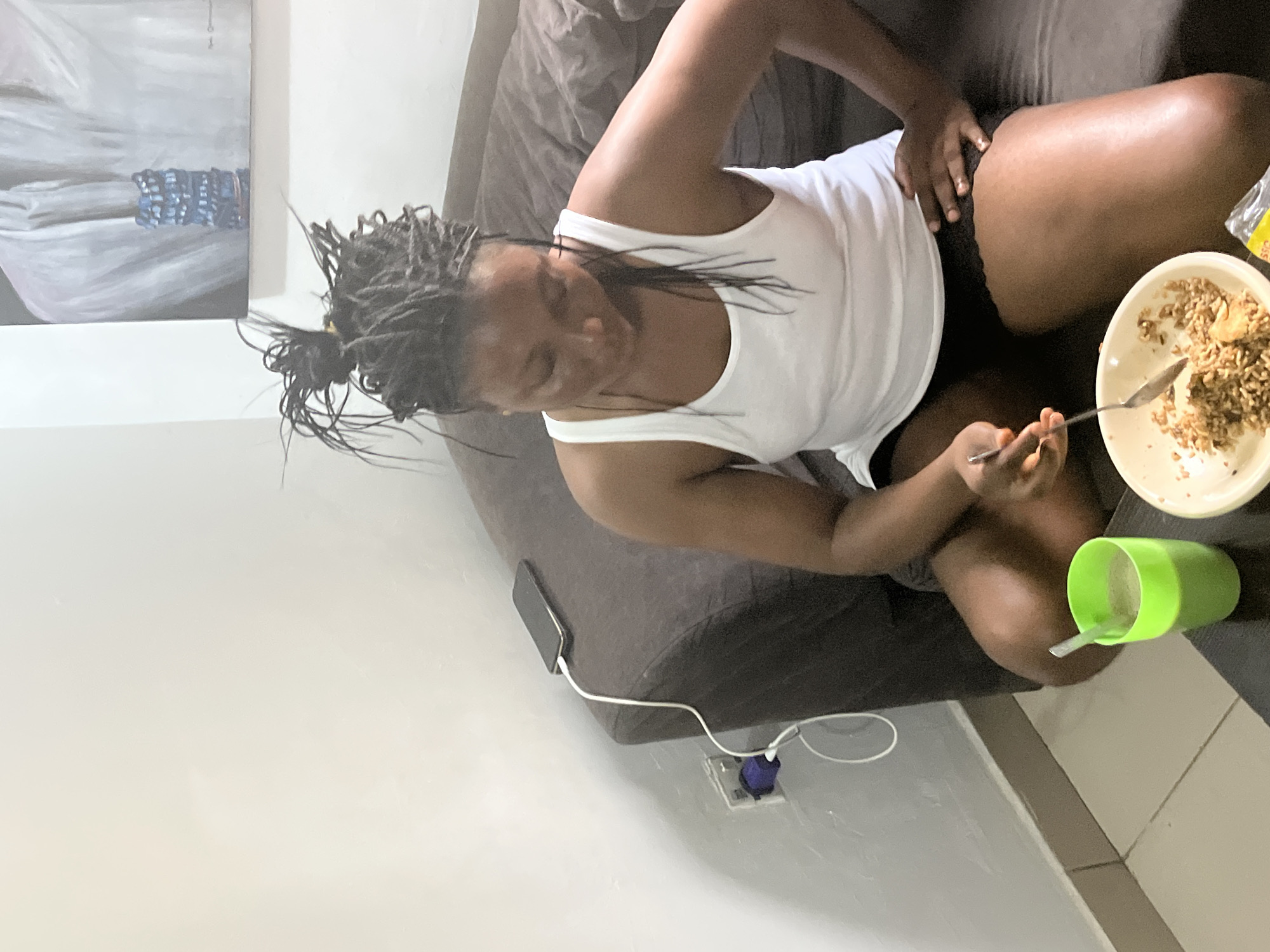 The image depicts a person sitting on a dark grey couch, seemingly indoors. The person is wearing a white tank top and dark shorts. They have dark, braided hair and appear to be looking down at a bowl of food in front of them, with a spoon in their hand. To their right, there is a bright green cup and a partially visible plastic bag. A cell phone is on the couch next to the person, plugged into a charger that is connected to a wall socket. In the background, there is a painting on the wall. The lighting suggests it could be daytime, but the exact time of day is not specified. The overall scene appears casual and relaxed. No text is visible in the image. The location context of Abuja, Nigeria is provided, but there are no specific visual cues in the image that directly indicate this location.