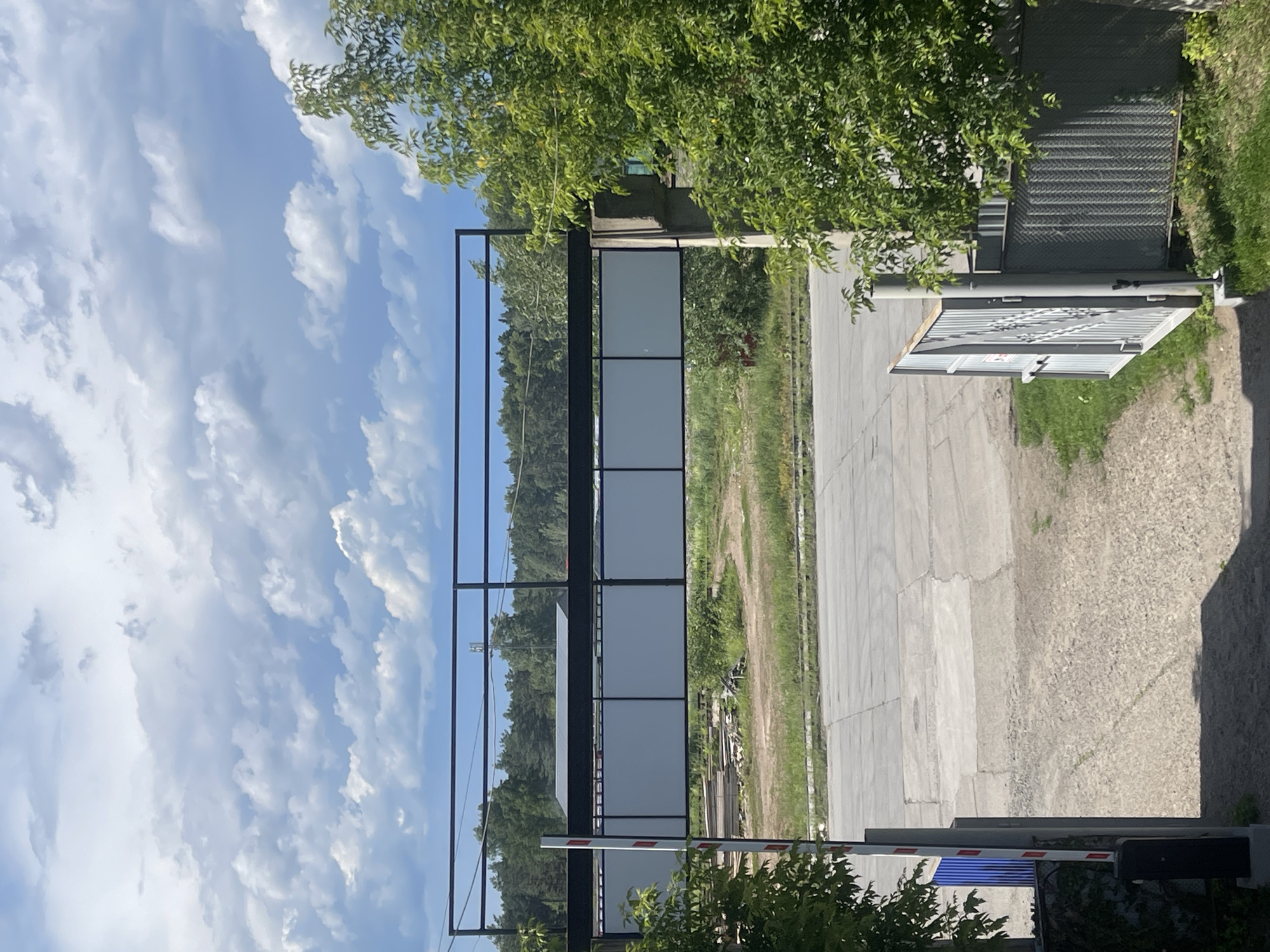 The image depicts an outdoor scene in Chaikovsky, Russia, under a cloudy sky. In the foreground, a gravel-covered pathway leads towards an open metal gate on the right and a white barrier with red stripes on the left, suggesting an entrance or checkpoint. Beyond the gate, a chain-link fence runs along the right edge of the frame, bordered by green grass and a large leafy tree. To the left, a portion of a blue fence is visible.
The middle ground features a paved area with distinct concrete slabs, likely a road or parking lot. Further back, a raised structure with a black metal railing and translucent white panels is visible, supported by concrete pillars. This structure appears to be elevated above a grassy area with scattered lumber and vegetation.
The background consists of a dense line of trees forming a forest. In the far distance, a cell tower can be seen rising above the treeline. The overall impression is that of an industrial or semi-industrial area adjacent to a natural landscape.
