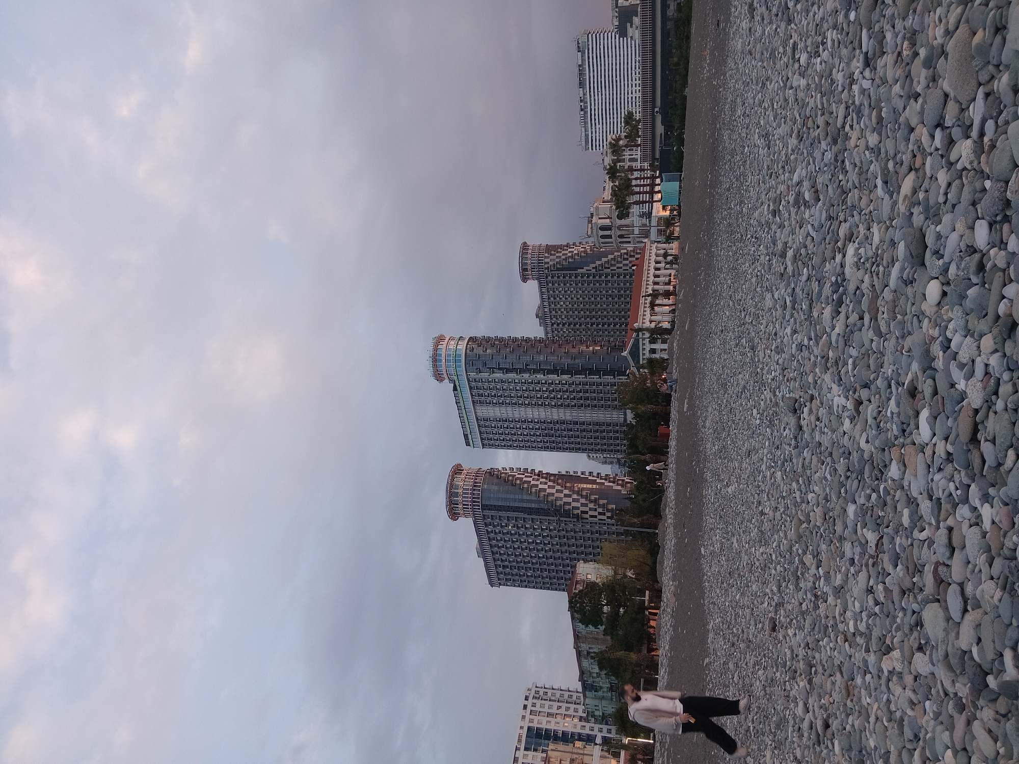 The image depicts a view of Batumi, Georgia, taken at dusk or dawn. The foreground is dominated by a rocky beach composed of smooth, rounded stones. In the mid-ground, several modern, high-rise buildings stand along the coastline. These buildings are notable for their contemporary architectural style, featuring rounded tops and grid-like facades. A person wearing a light-colored jacket and dark pants is walking on the beach. The sky is overcast with pale clouds.